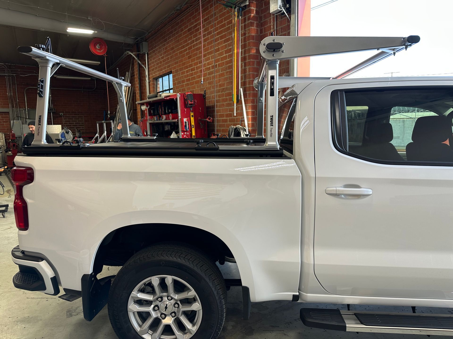 A white pickup truck with an adjustable, silver metal rack system installed over the truck bed in a garage workshop.