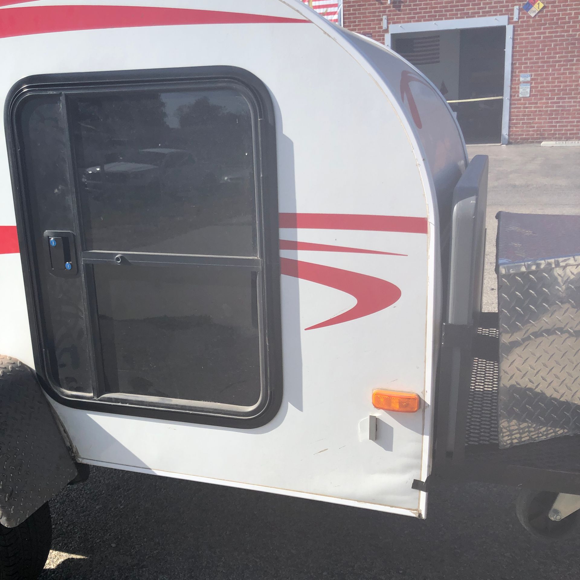 Side view of a white teardrop camper trailer with red accents and a black door, parked on a paved lot.