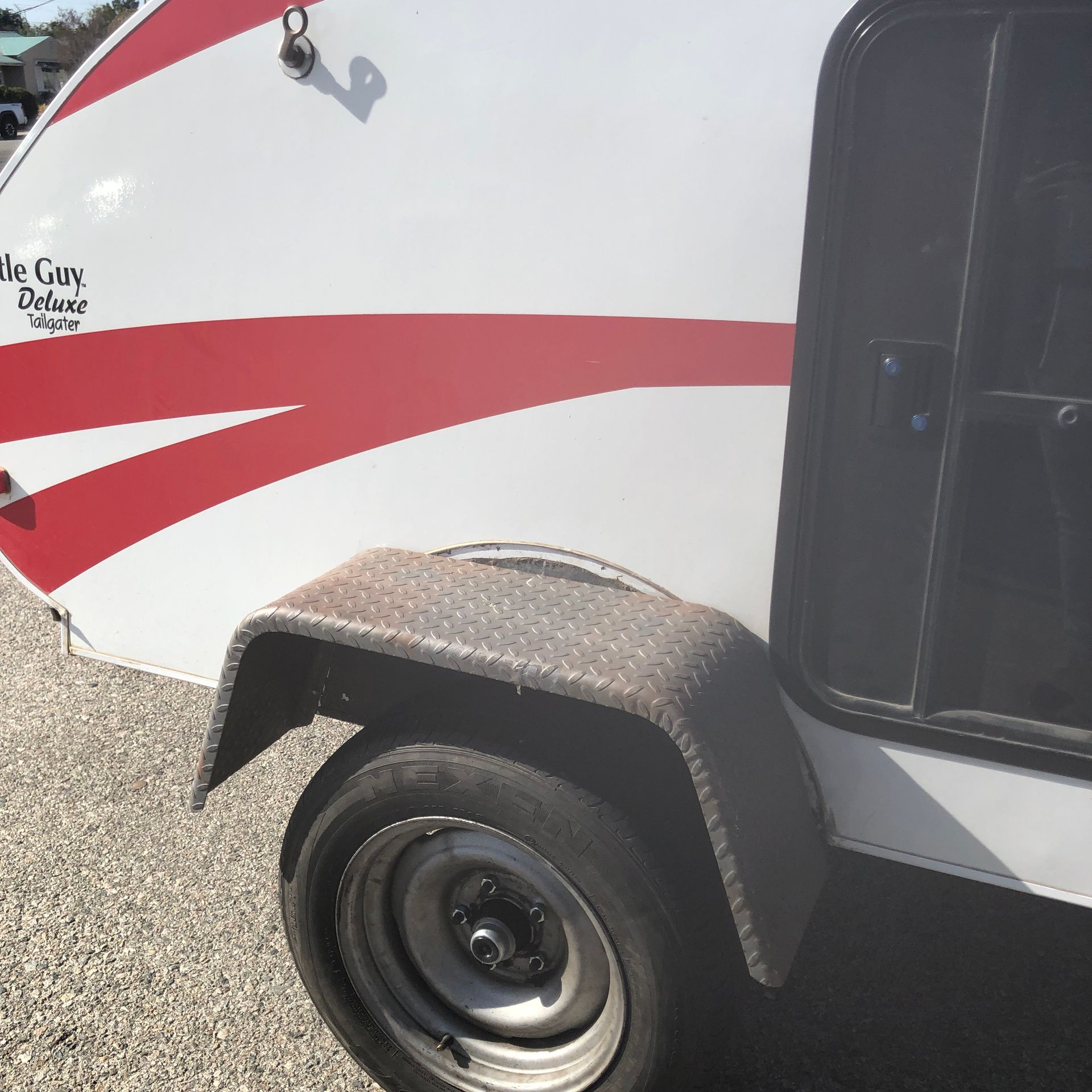 A white teardrop camper with red stripes and a diamond-plate metal fender over a black tire on a gravel surface.