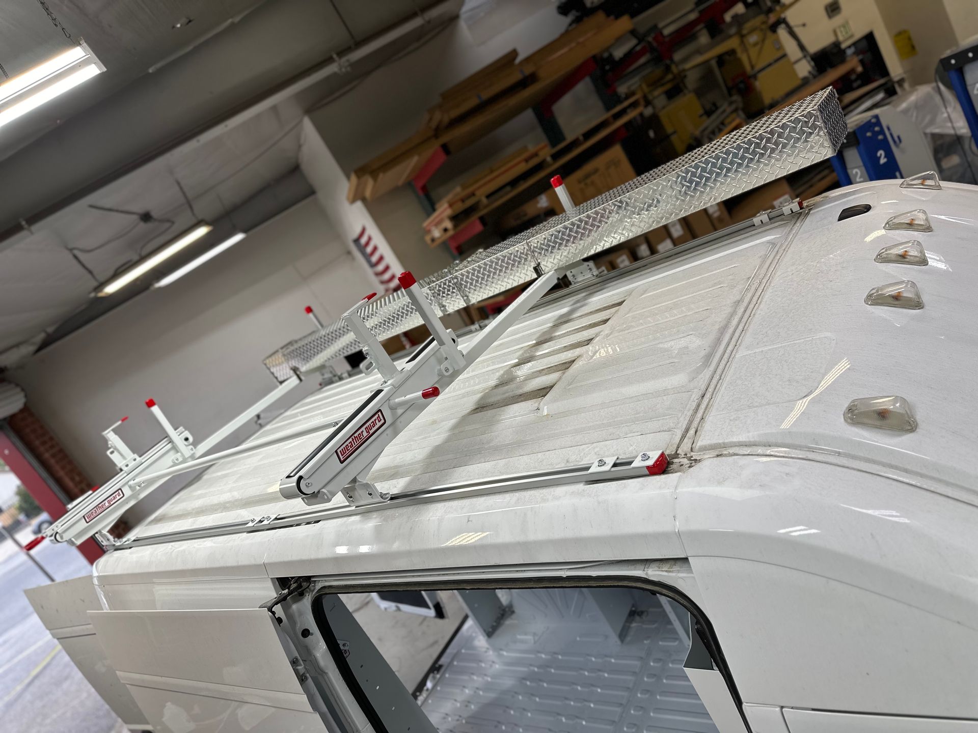 White van with a ladder rack system mounted on the roof, positioned in an indoor workshop or warehouse setting.