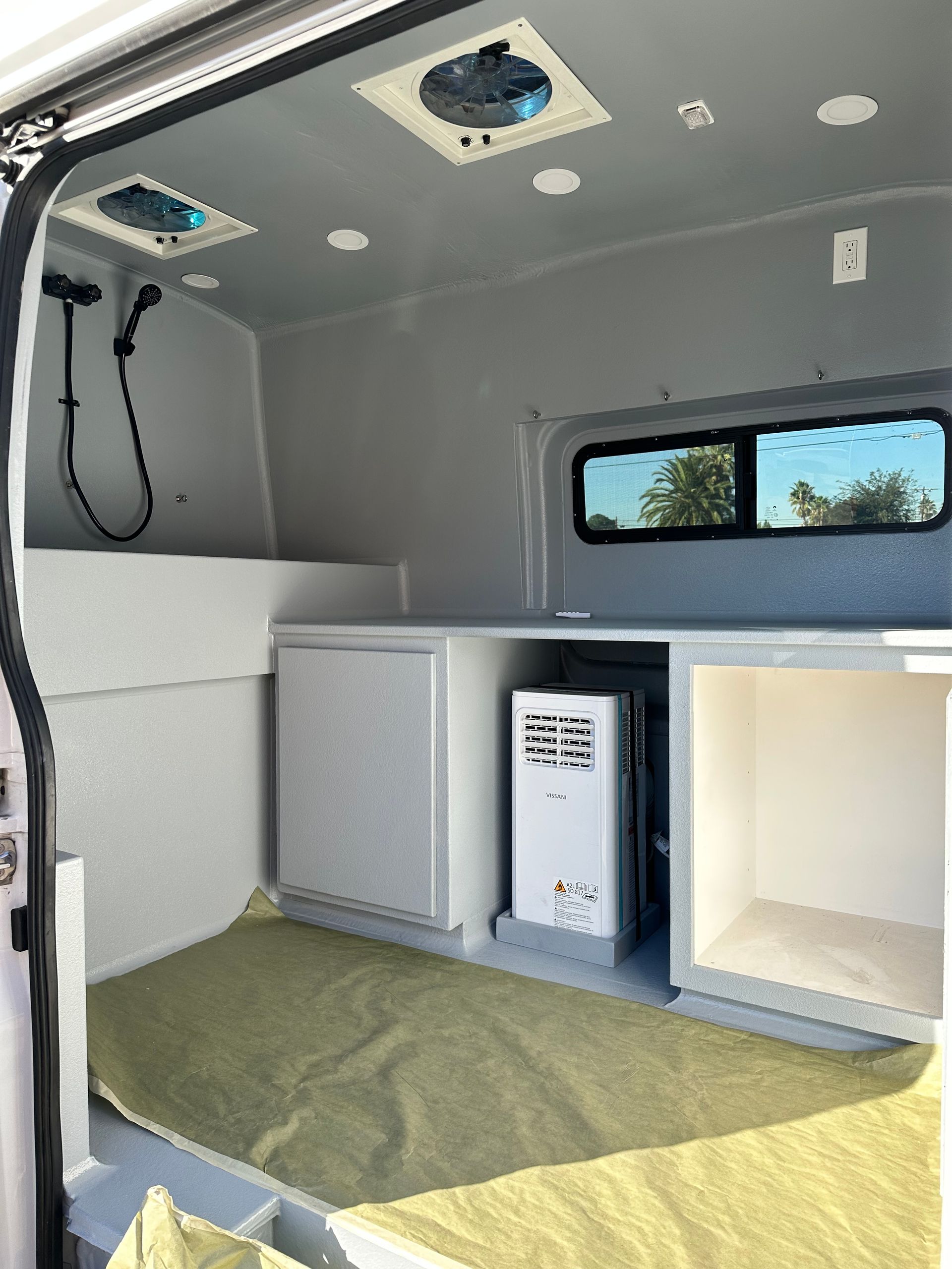 Interior of a van conversion featuring gray walls, two ceiling vents, a shower head, a cabinet, and a water heater.