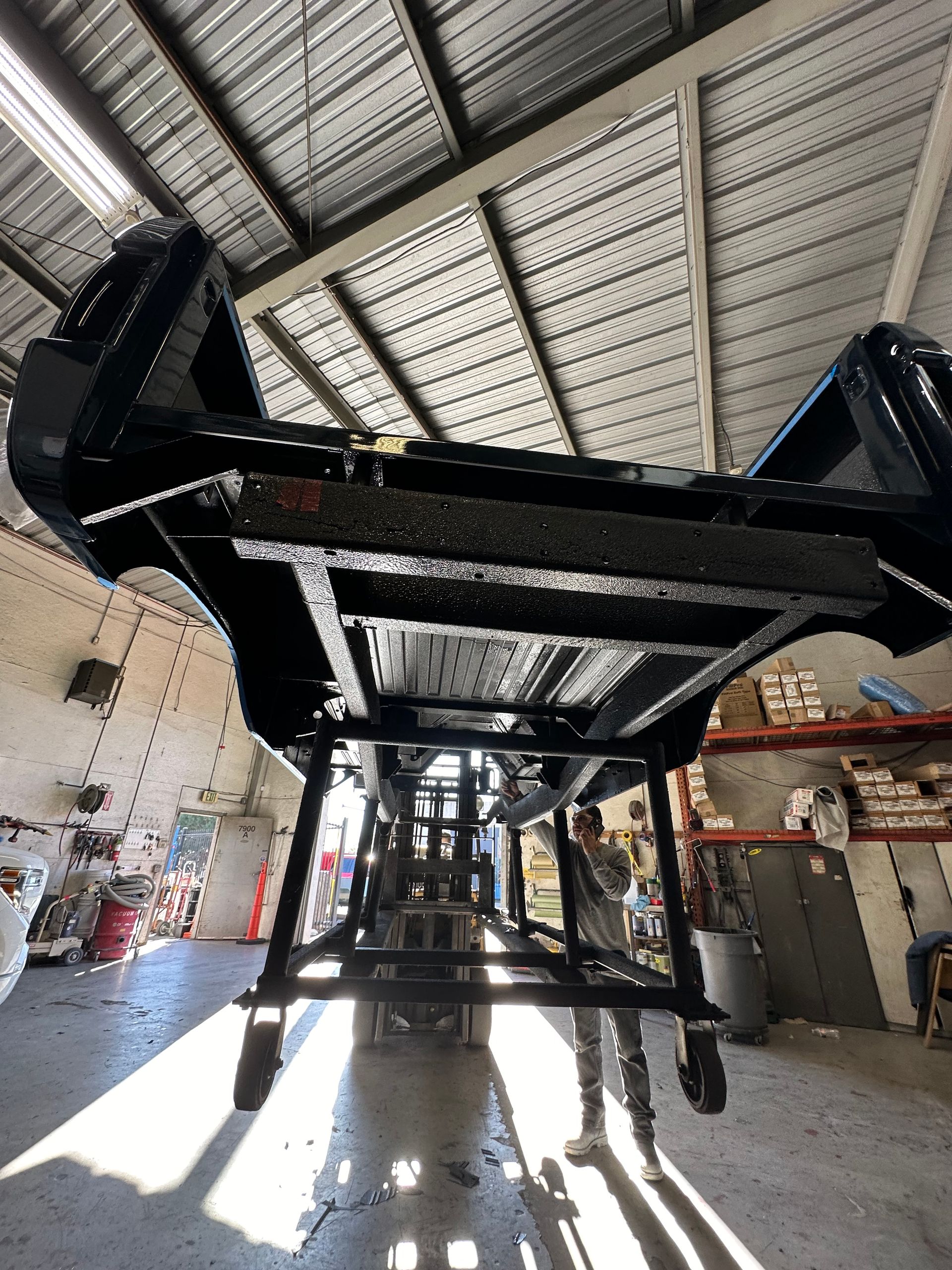 A person stands beneath a black vehicle chassis mounted on a rotating metal frame inside an automotive workshop.