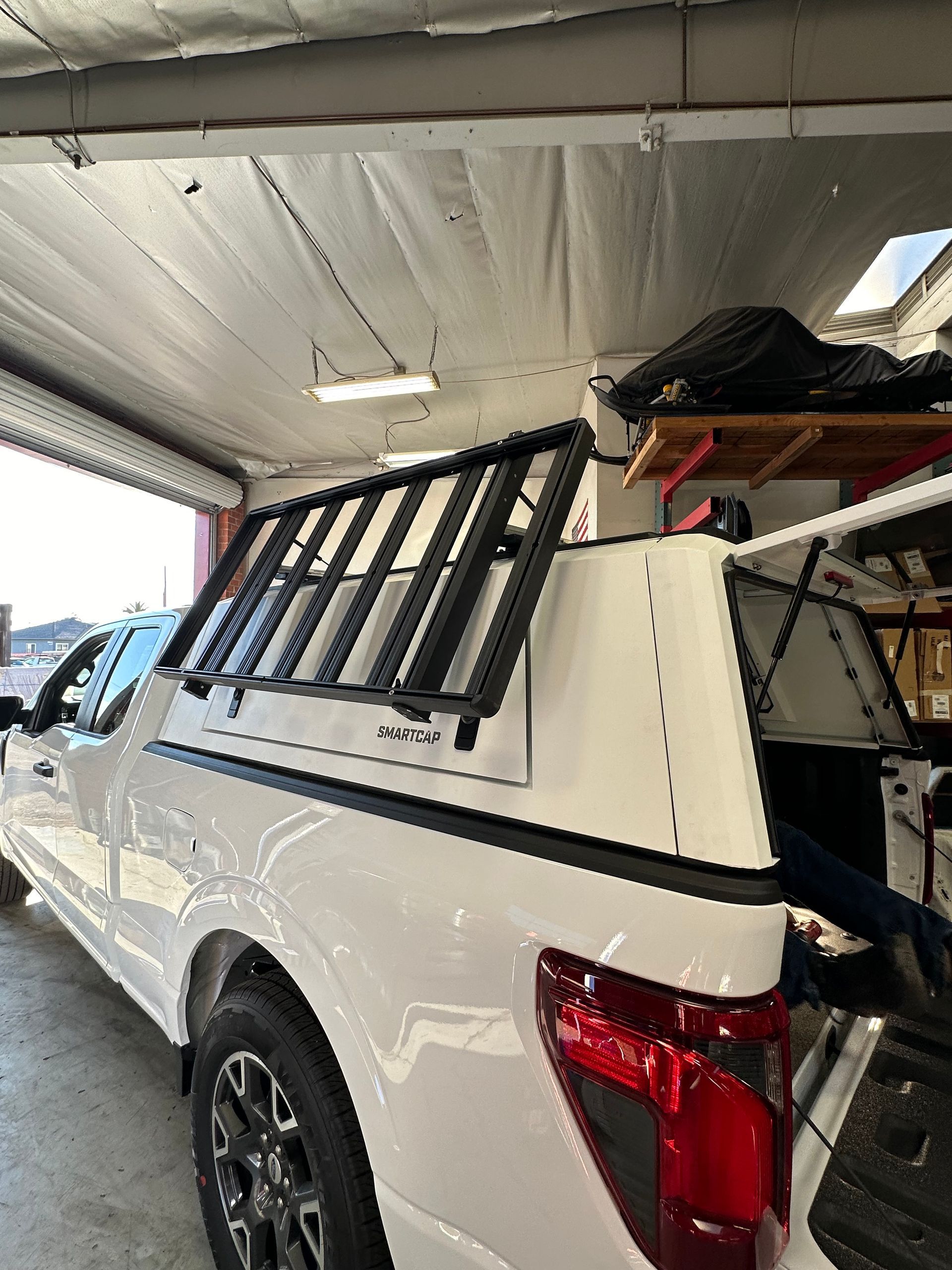 A white pickup truck with a white camper shell featuring a black roof rack mounted on its side in a garage.