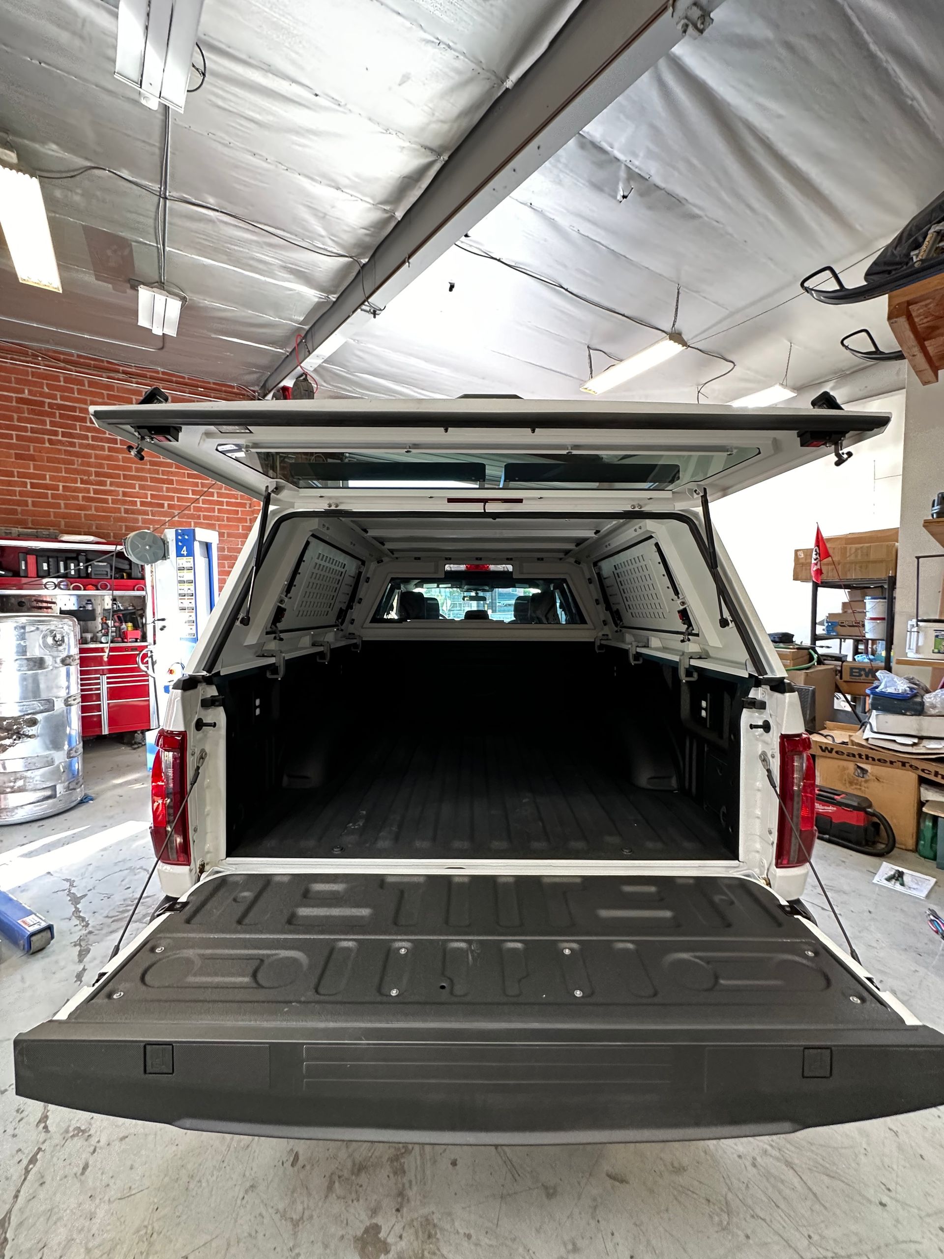 The open tailgate and bed of a white pickup truck, equipped with a matching white camper shell in a garage setting.