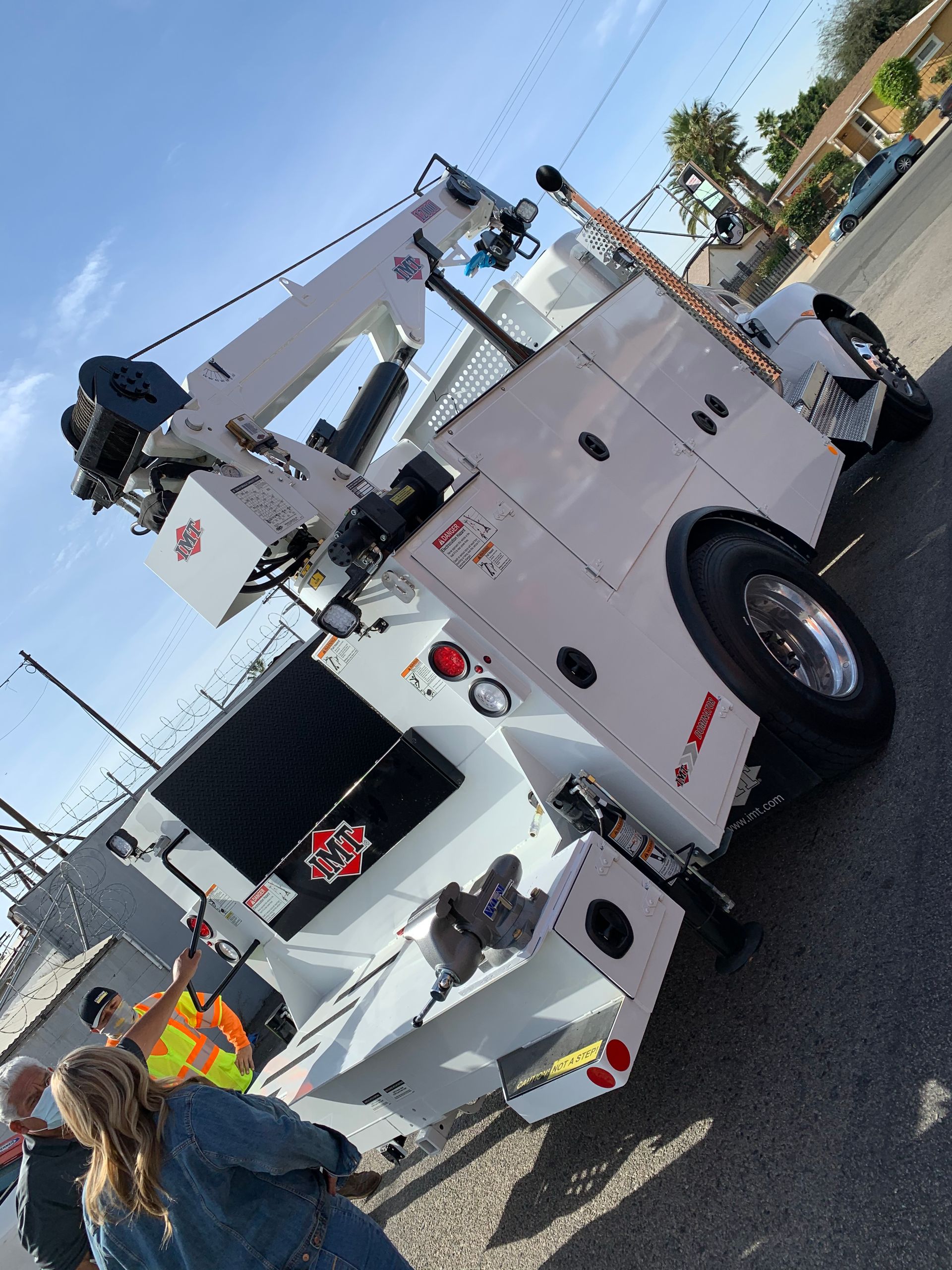 A white service truck with a hydraulic crane is parked outdoors on an asphalt lot near a group of people.