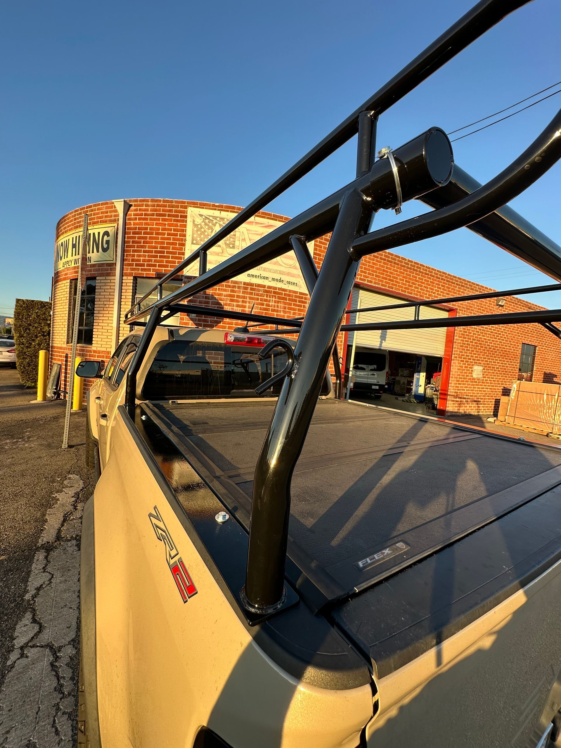 A beige pickup truck with a black overhead utility rack parked outside a building with orange-and-white corrugated siding.
