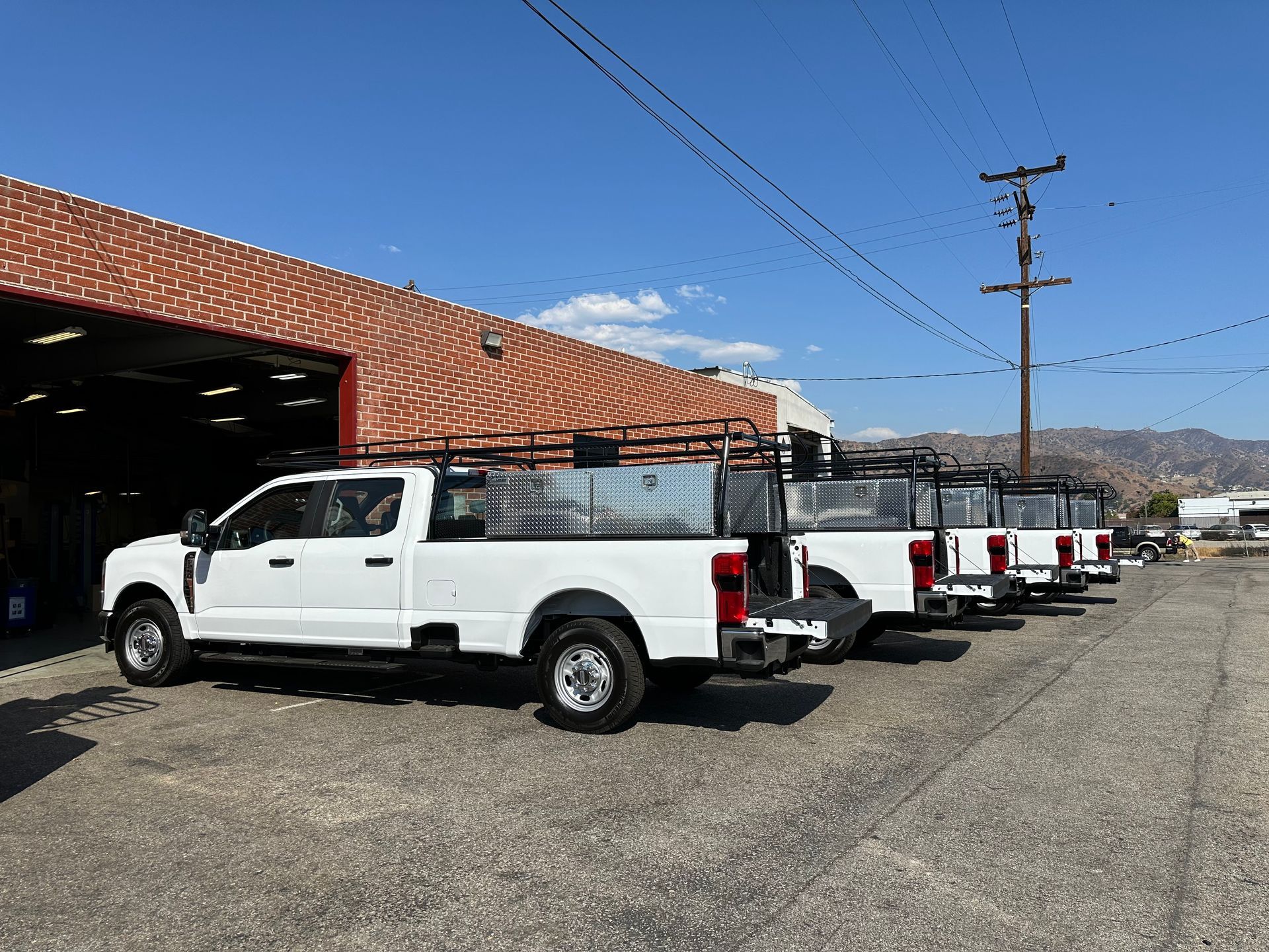 A line of white pickup trucks with metal utility racks parked in a gravel lot outside a brick building under a blue sky.