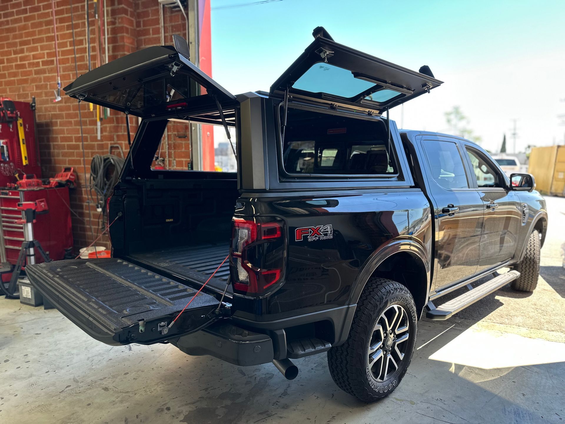 A black pickup truck parked indoors with its tailgate and camper shell side panels open, revealing the empty truck bed.