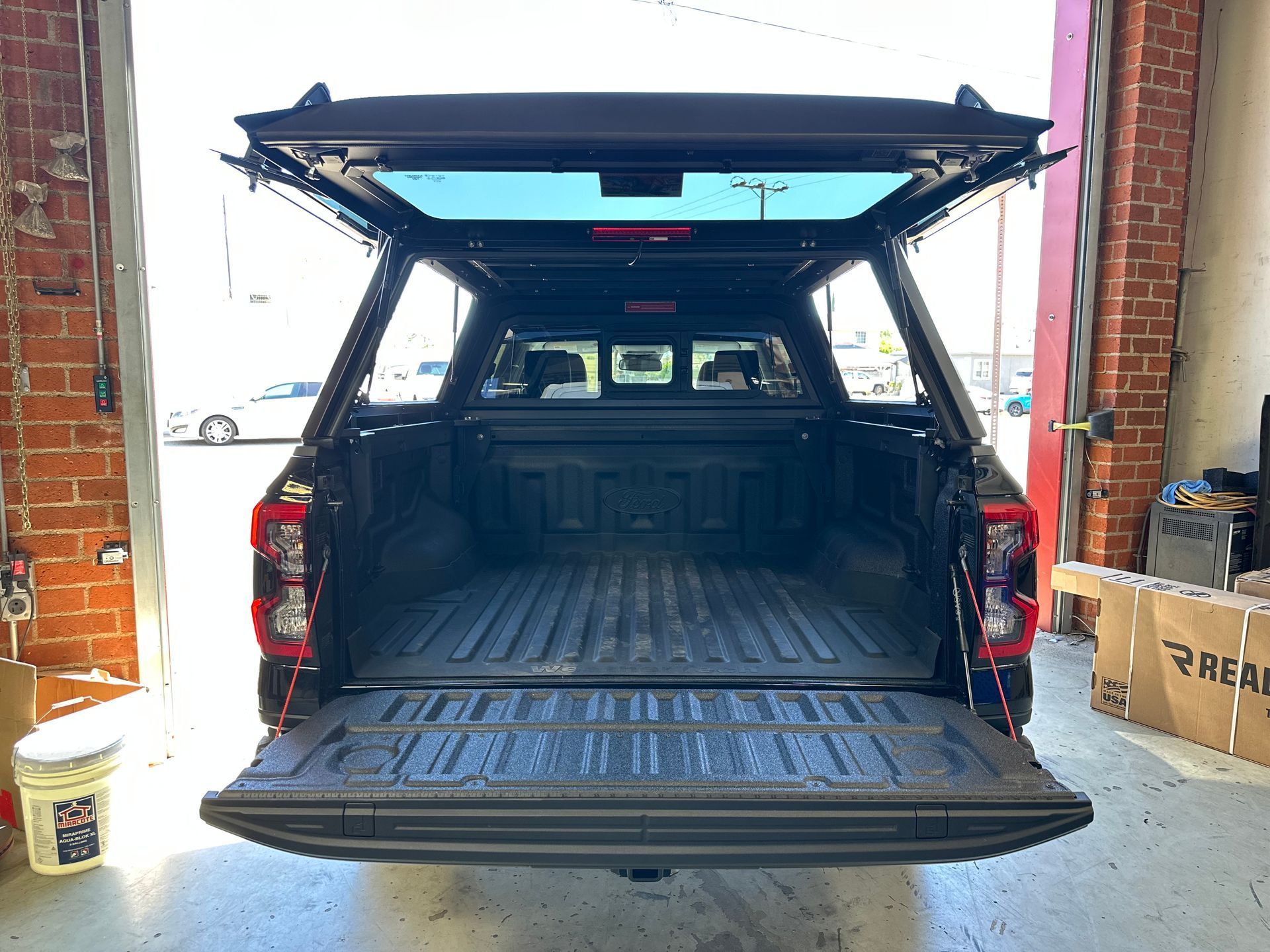 Rear view of a black pickup truck with its tailgate and fiberglass canopy topper door open in a workshop.