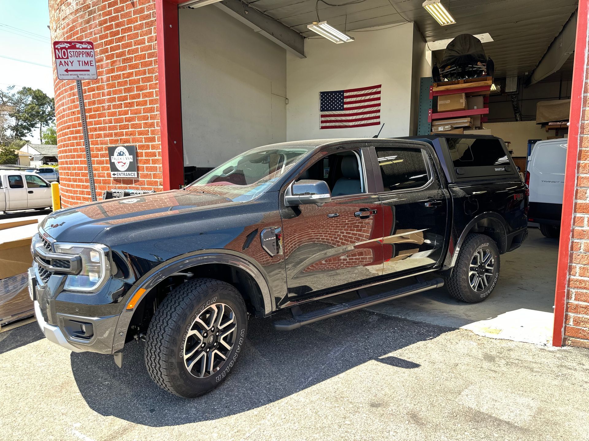 A black Ford Ranger pickup truck with a camper shell parked in front of an automotive shop with a red brick exterior.