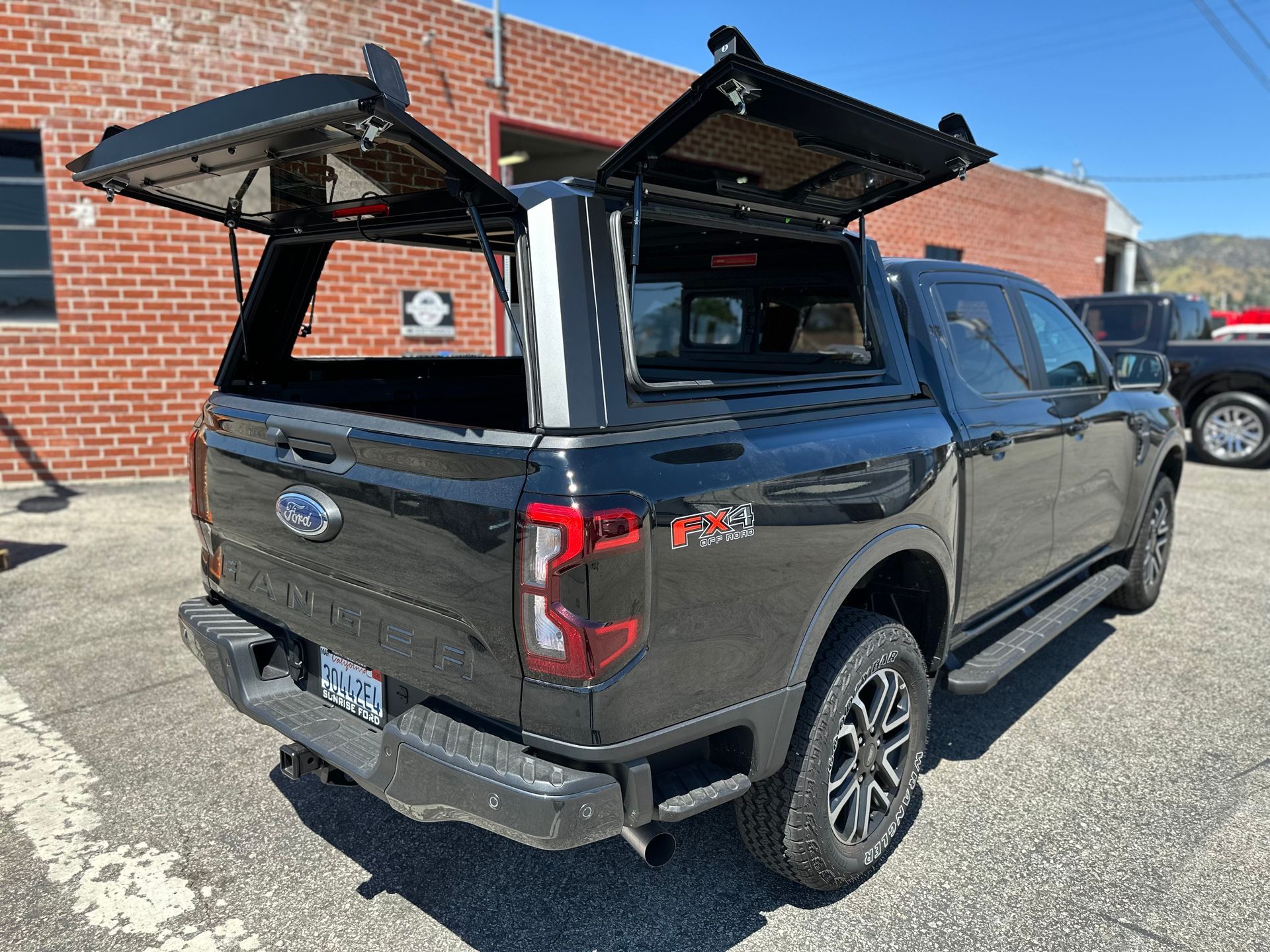 Black Ford pickup truck with a black camper shell, rear and side access hatches open, parked outside a brick building.