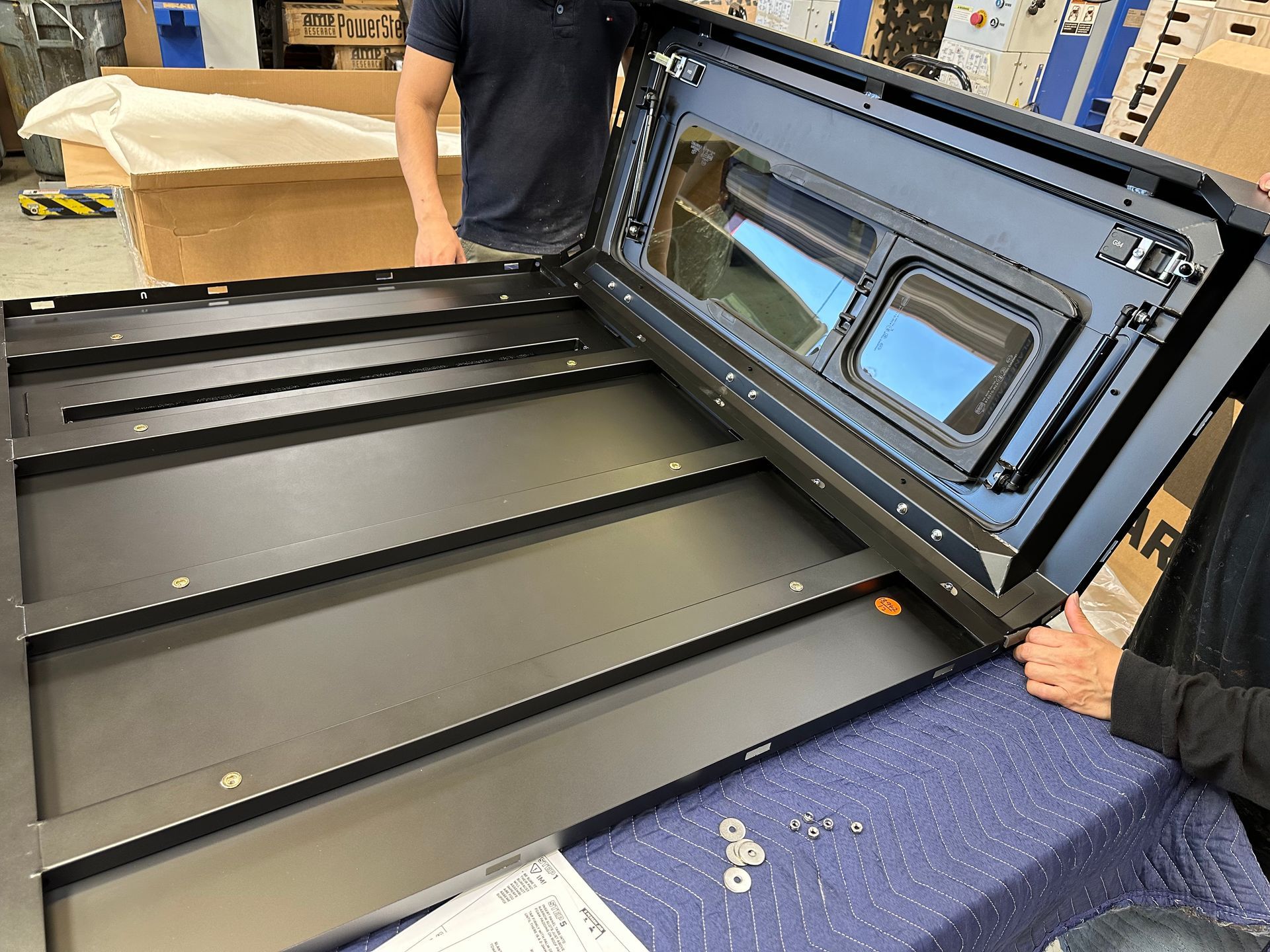 Two people assemble a large, black metal truck bed cover on a workbench in a workshop.