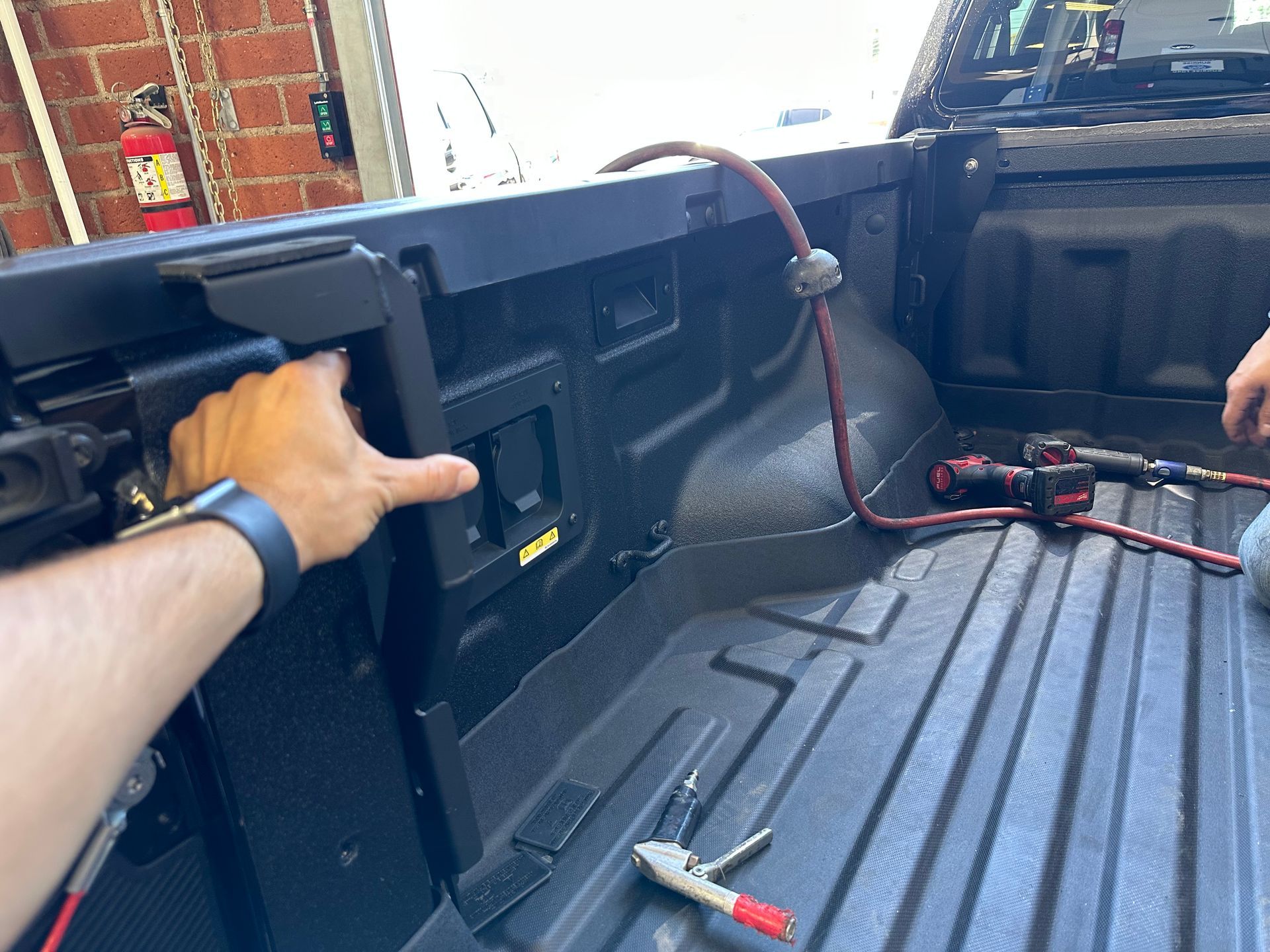 A person installing a black metal bracket in the bed of a pickup truck, with tools lying on the ribbed surface.