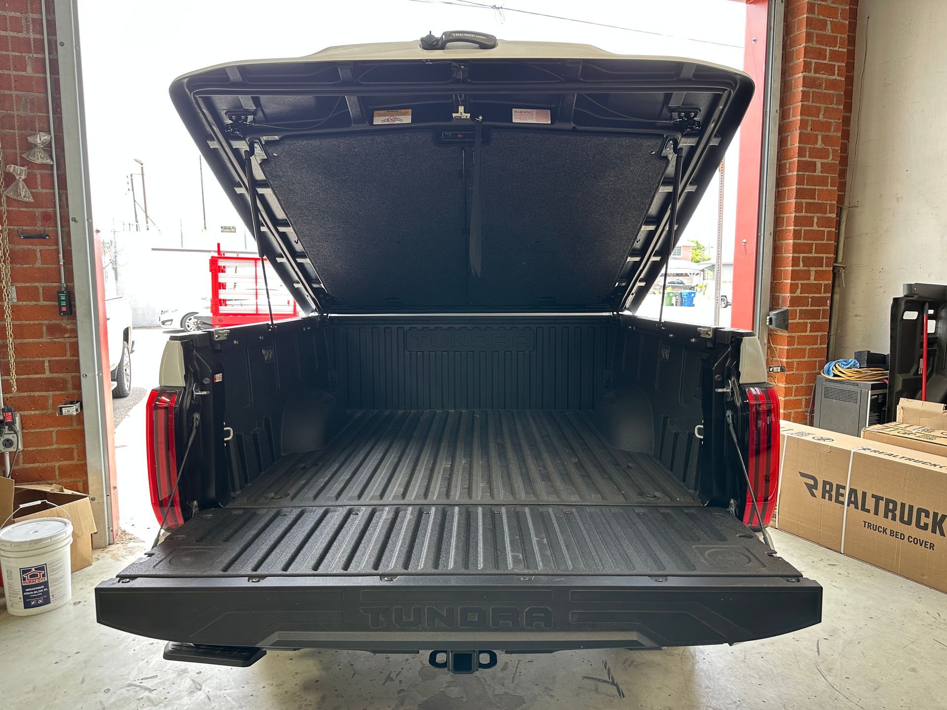 Open truck bed with a black tonneau cover and tailgate lowered, parked inside a garage with brick walls.