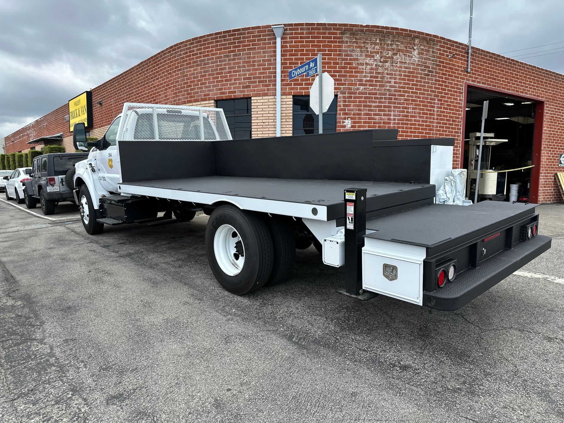 A white commercial flatbed truck parked on an asphalt lot in front of a red brick building.