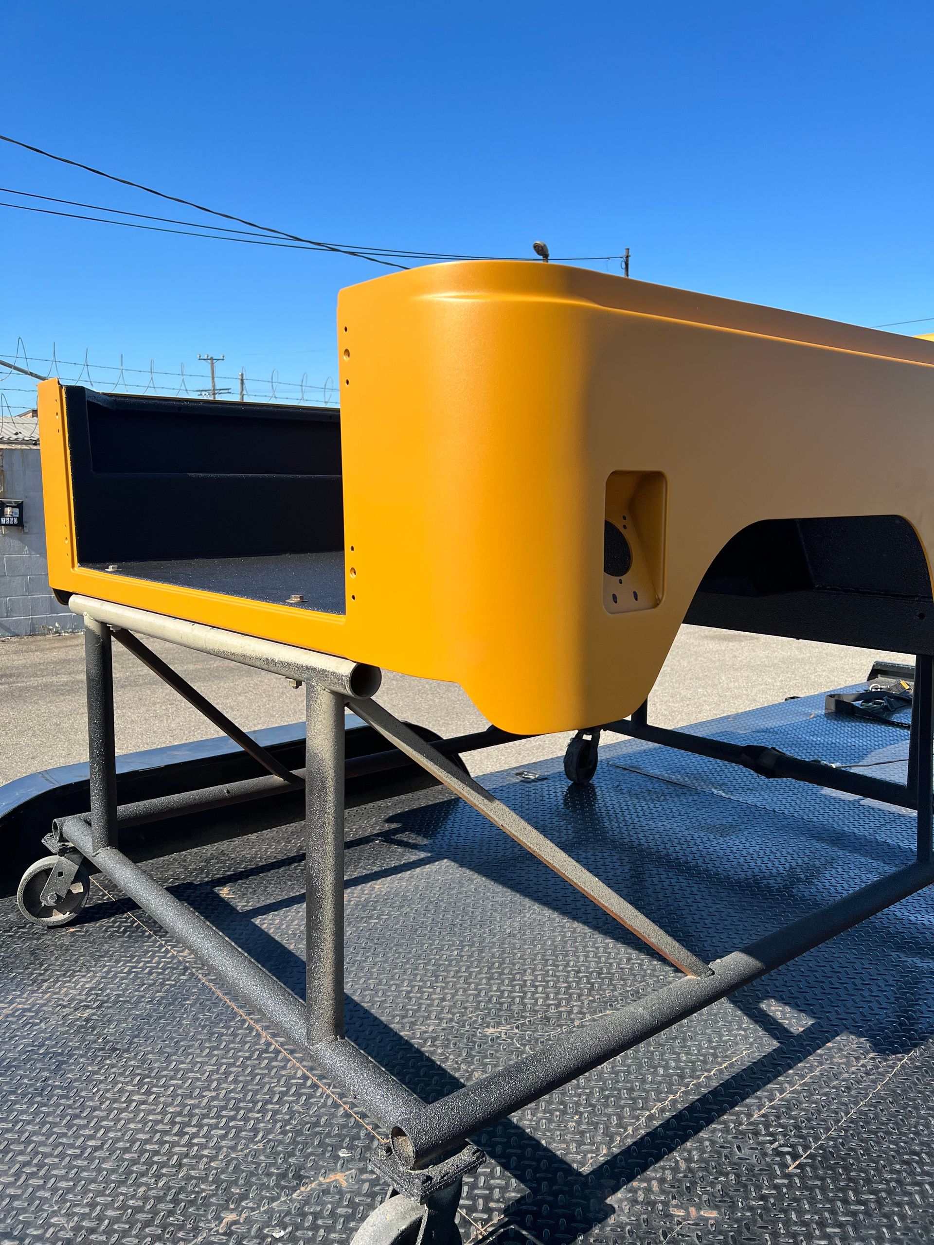 A mustard-yellow Jeep body tub rests on a metal rolling cart outdoors on a sunny day.