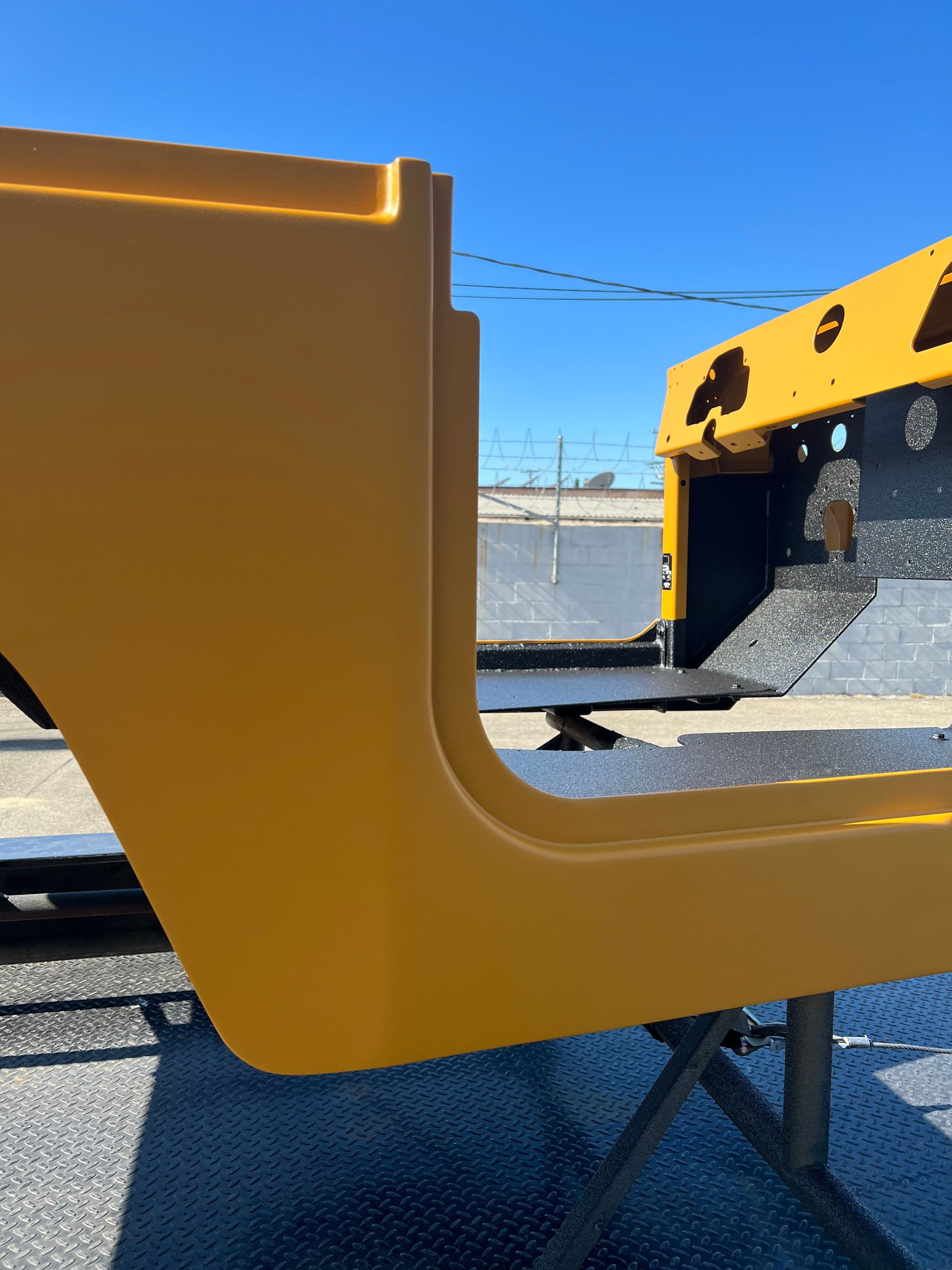 A bright yellow, unpainted jeep body tub sits on a metal stand outdoors against a clear blue sky.