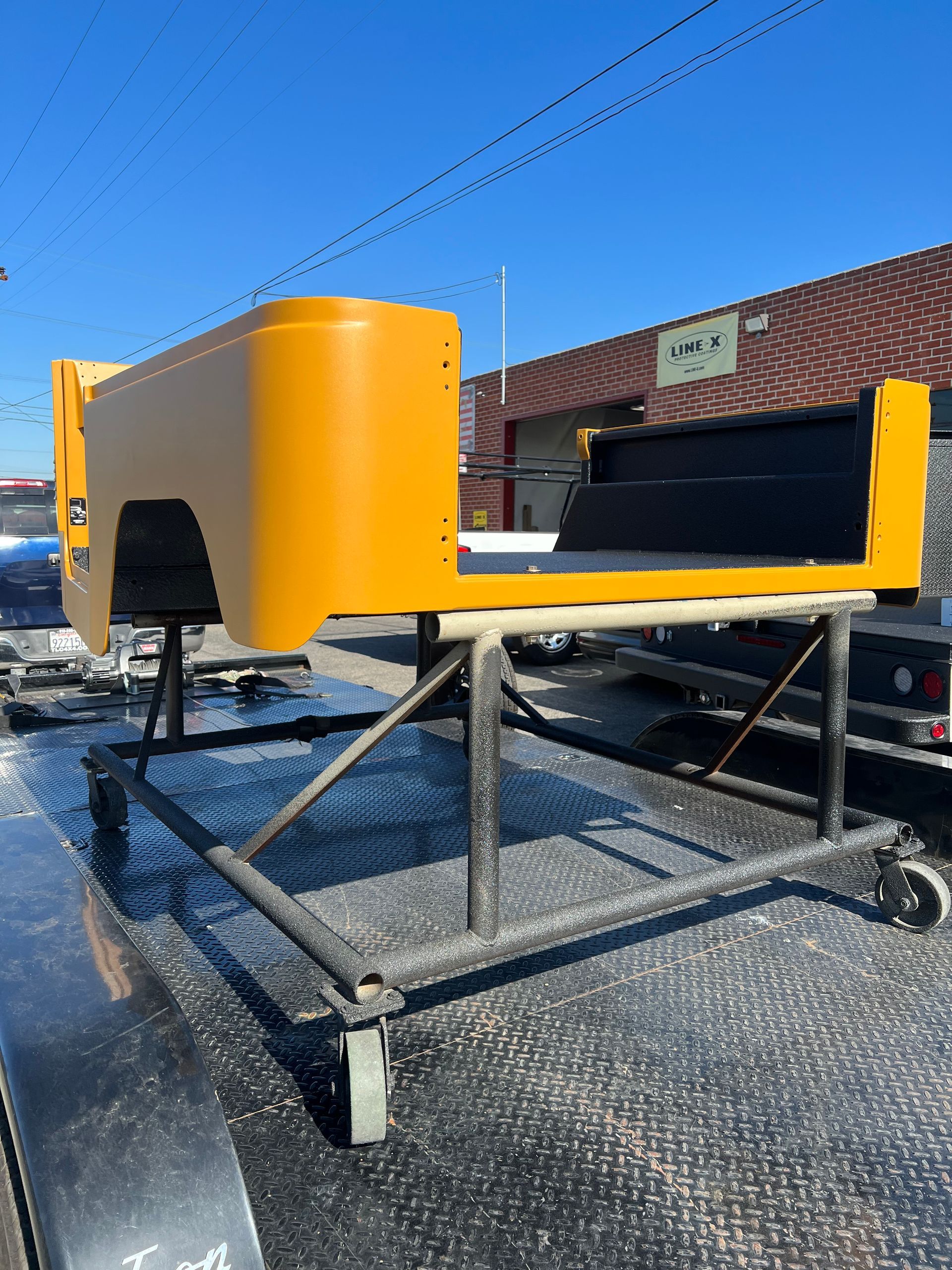 A yellow Jeep tub mounted on a black metal rolling cart, parked on a trailer in front of a brick building.