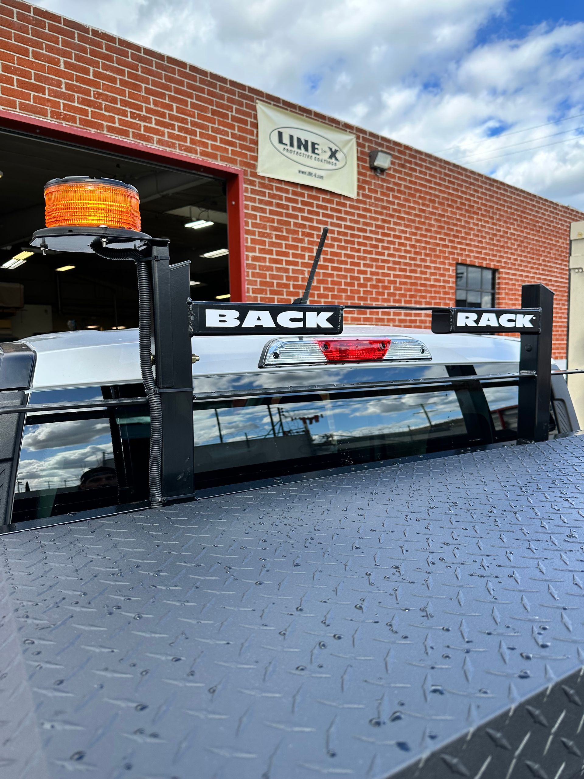 Black truck rack with an orange warning beacon installed on the bed of a pickup, parked outside a brick building.