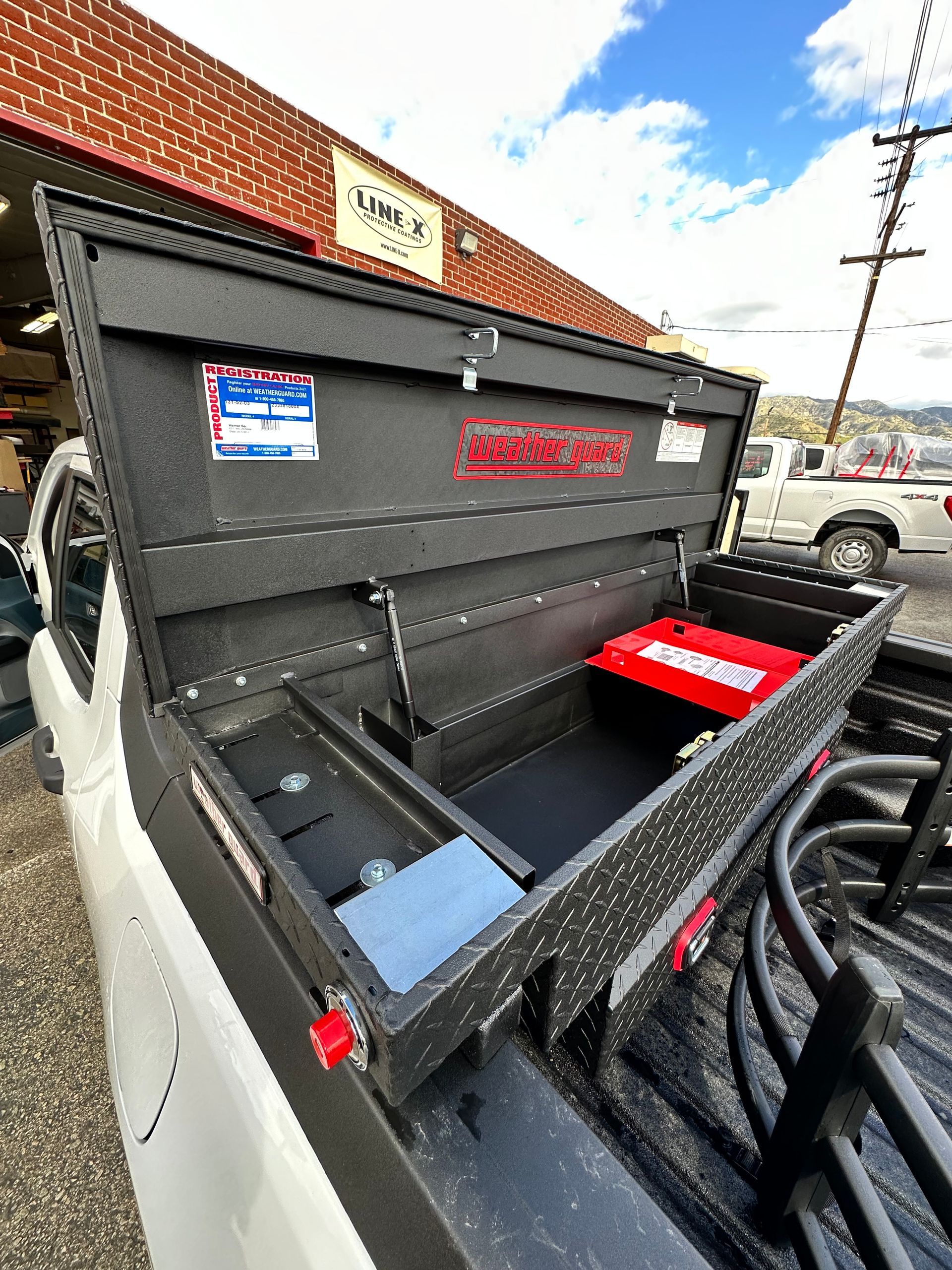 An open, black textured truck bed storage box with a red internal container, mounted on a white truck.