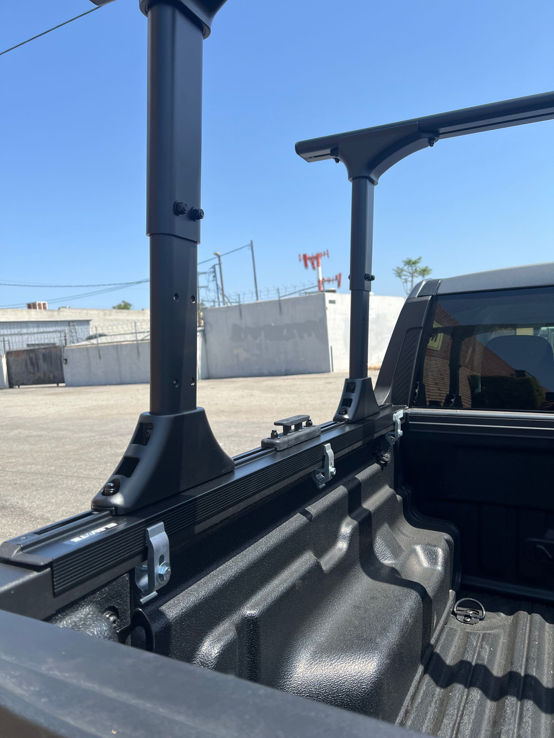 Black truck bed rack system with adjustable vertical supports mounted on a textured bed rail against a blue sky.