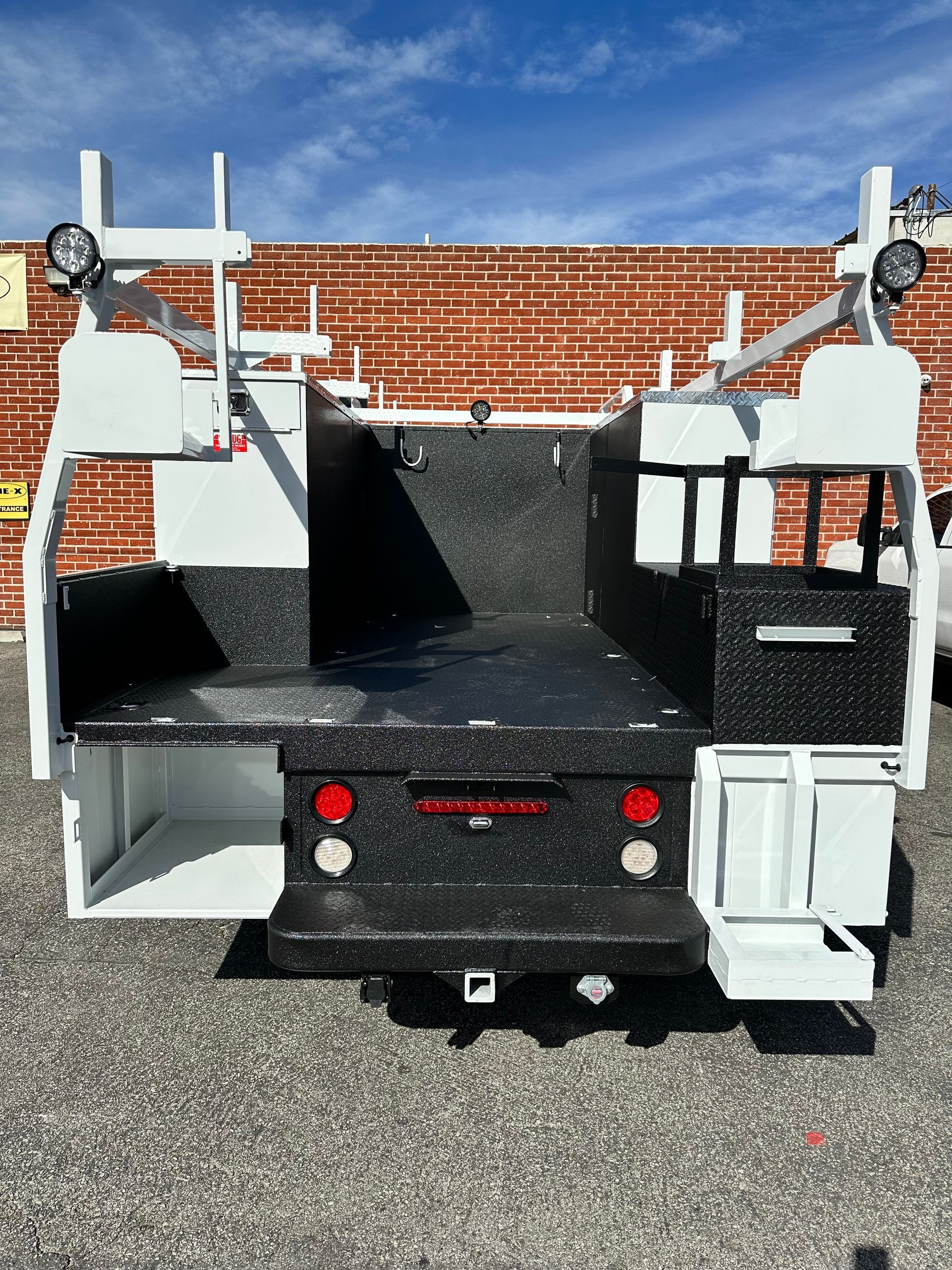 A white utility truck bed with a textured black coating on the floor and sides, featuring storage compartments and lights.