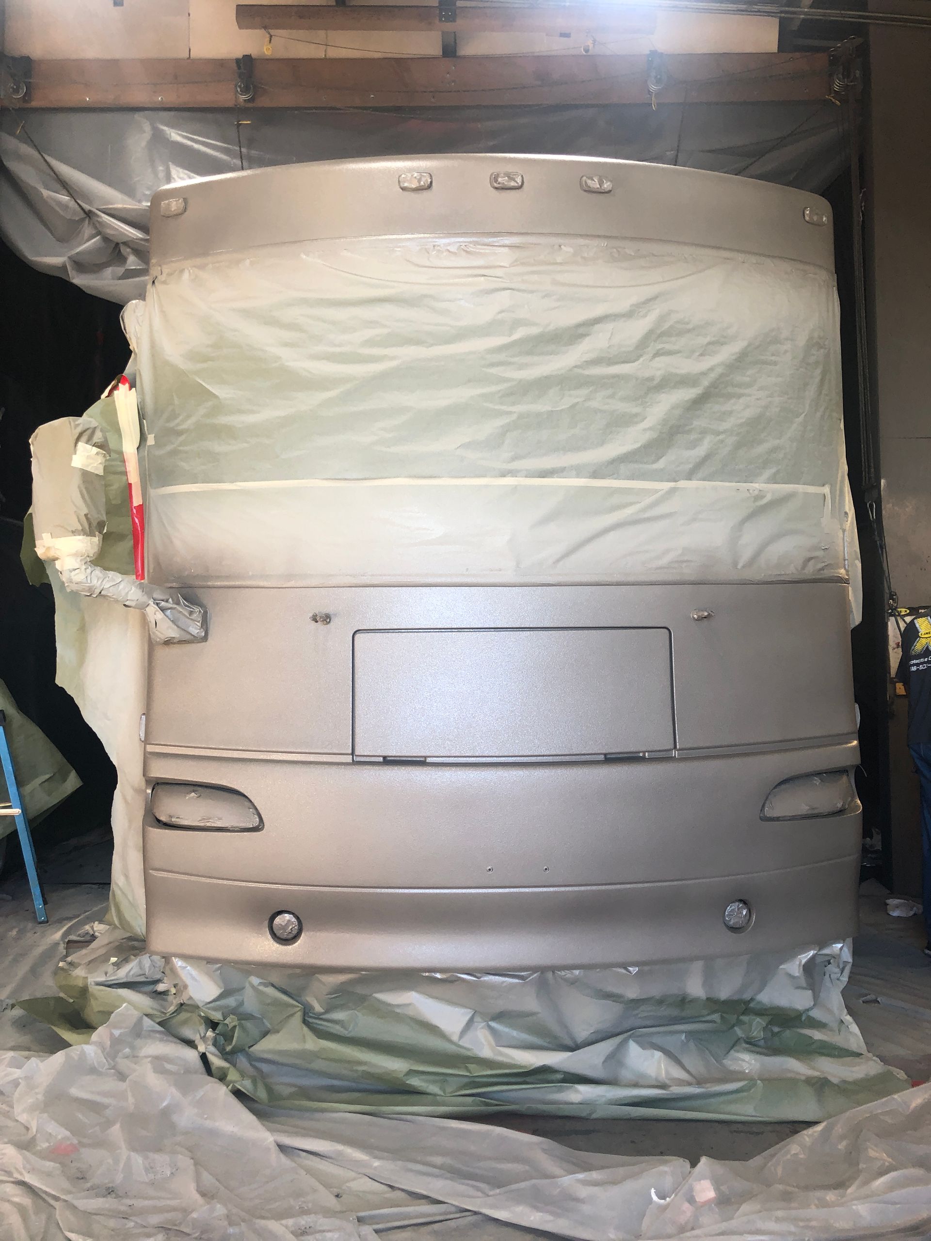 A silver-colored bus front is masked with protective plastic in a workshop, undergoing paint or repair.