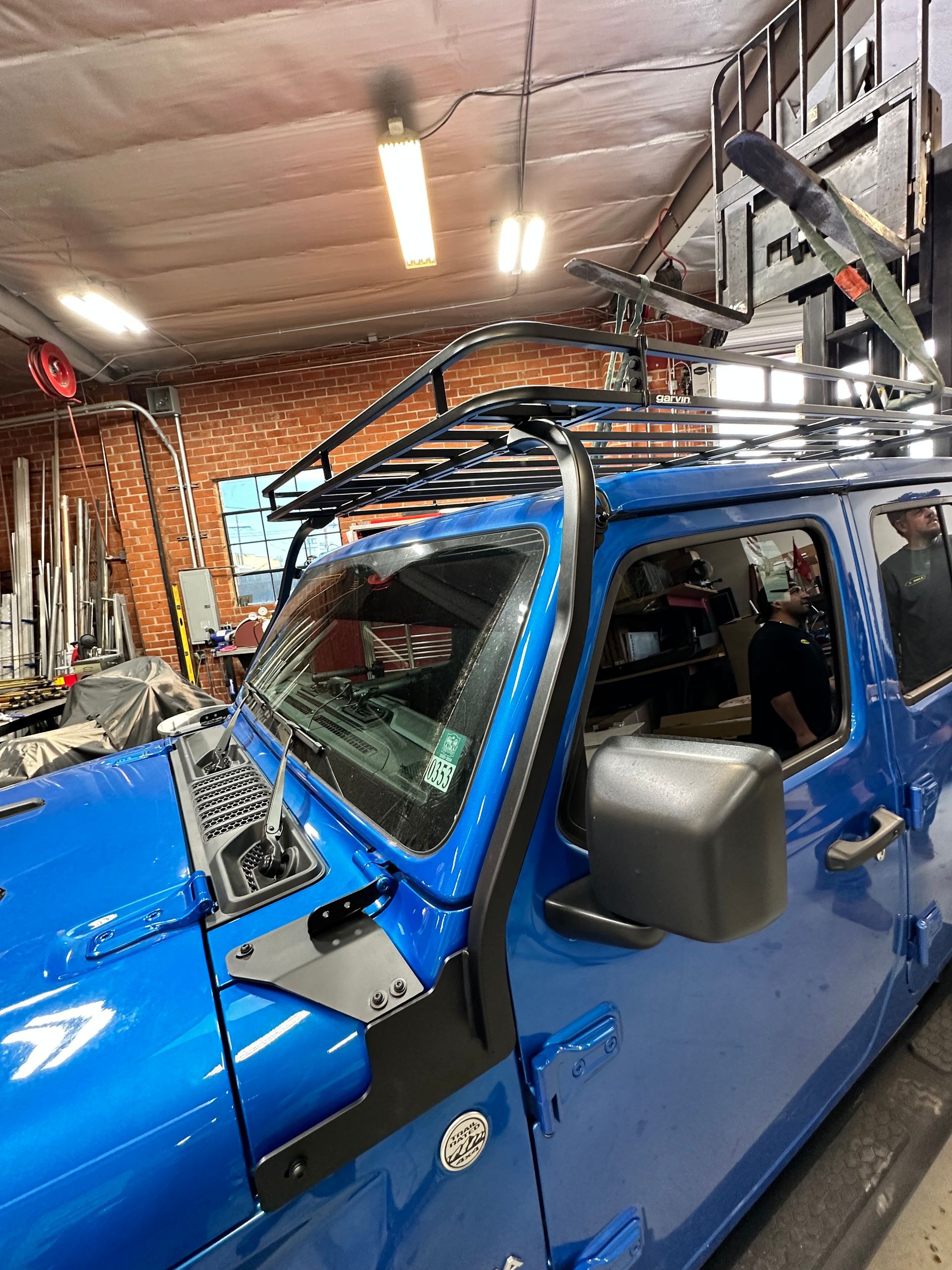A bright blue Jeep with a heavy-duty roof rack and an external A-pillar frame parked inside a brightly lit garage.