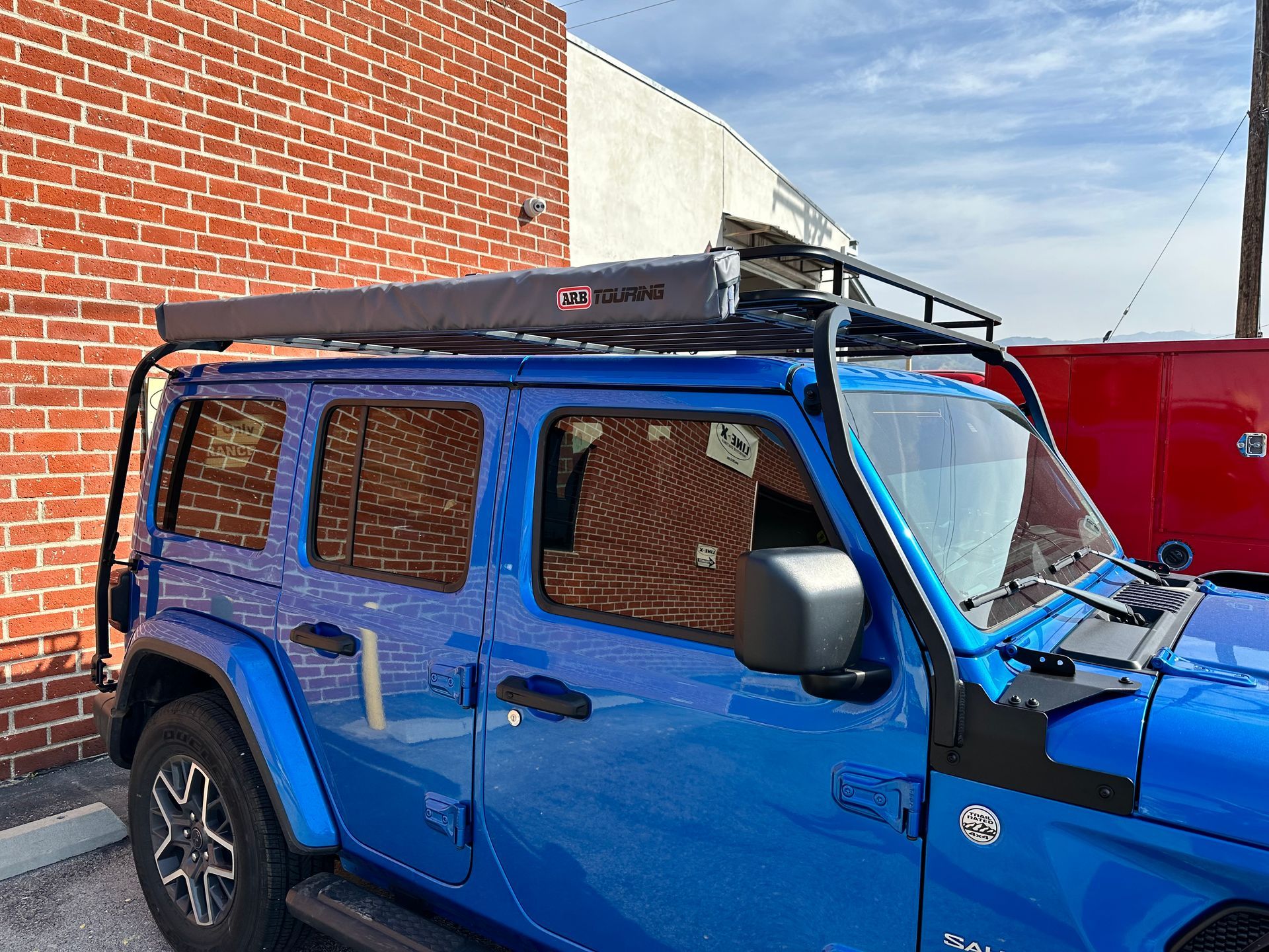 A bright blue Jeep Wrangler parked in front of a brick wall, equipped with a roof rack and a grey side-mounted awning.