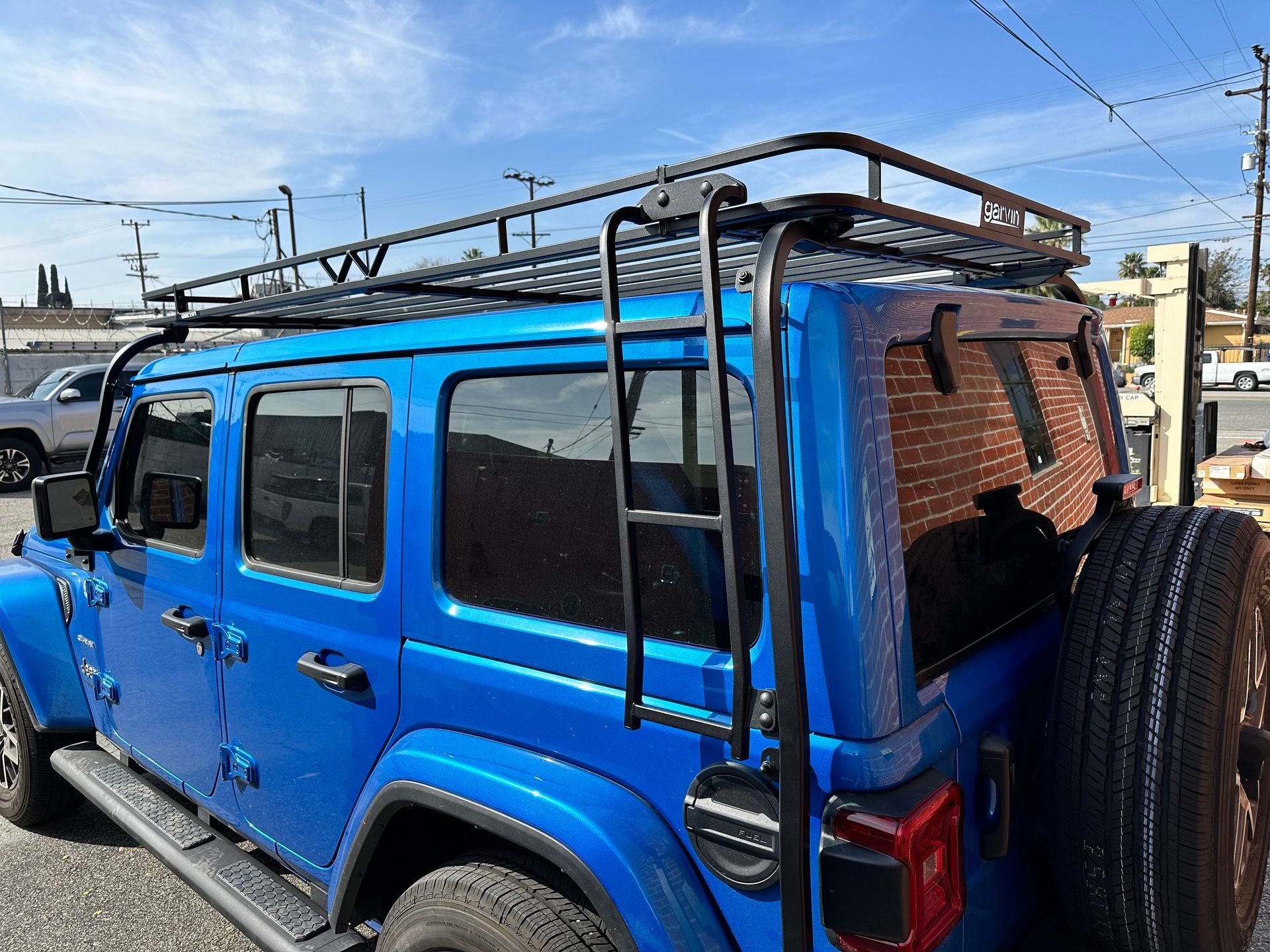 A bright blue Jeep SUV equipped with a black metal roof rack and rear-mounted ladder parked on a gravel lot.