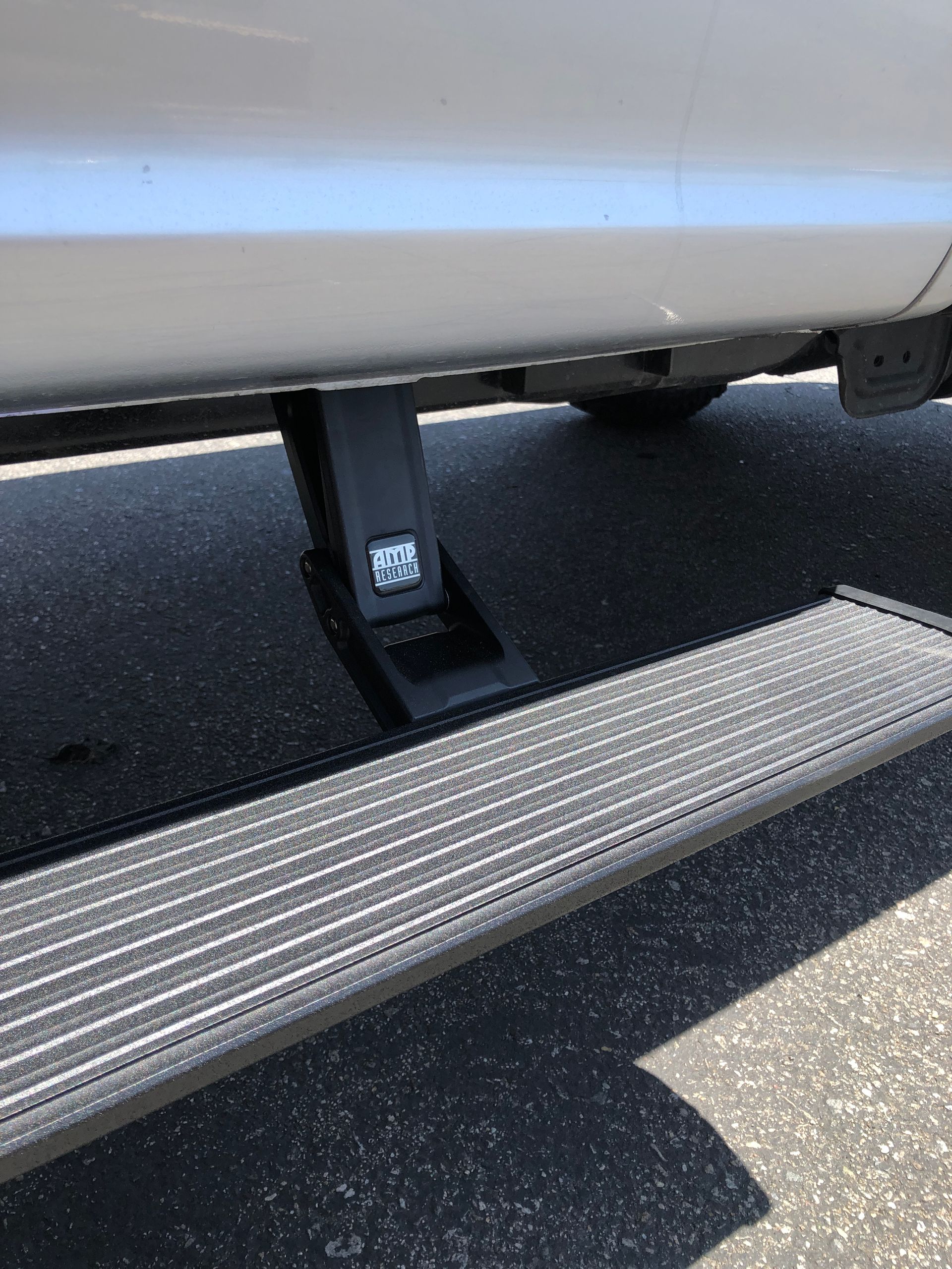 A black power-operated retractable running board extended from underneath a white vehicle over asphalt.