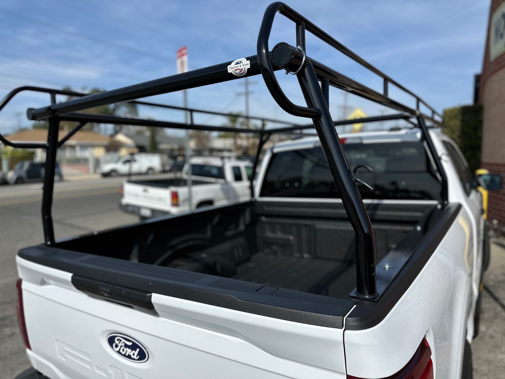 A black metal ladder rack installed on the bed of a white Ford pickup truck parked on a street.