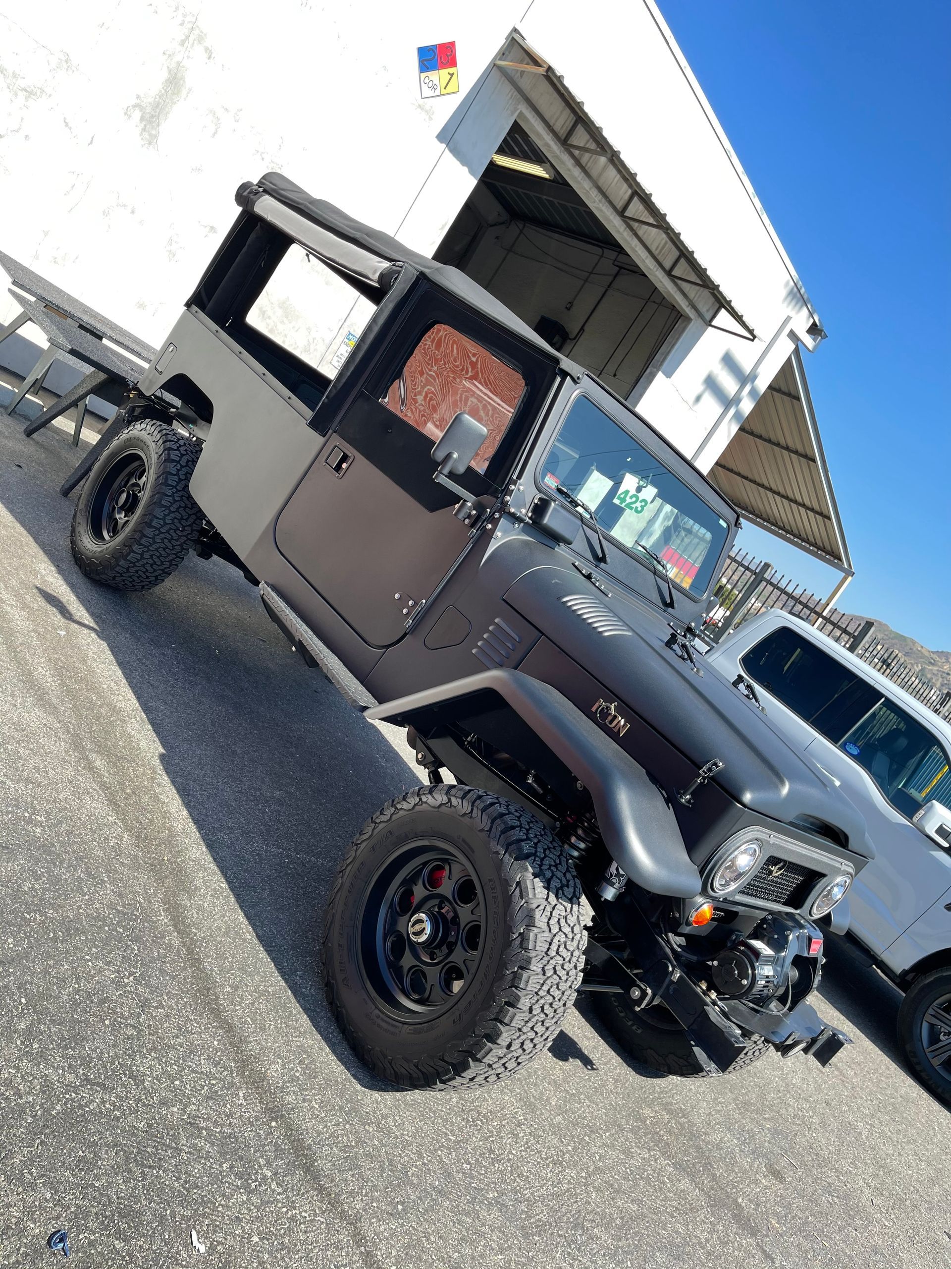 A dark gray, modified off-road vehicle with black wheels and a soft top parked on an asphalt lot near a building.