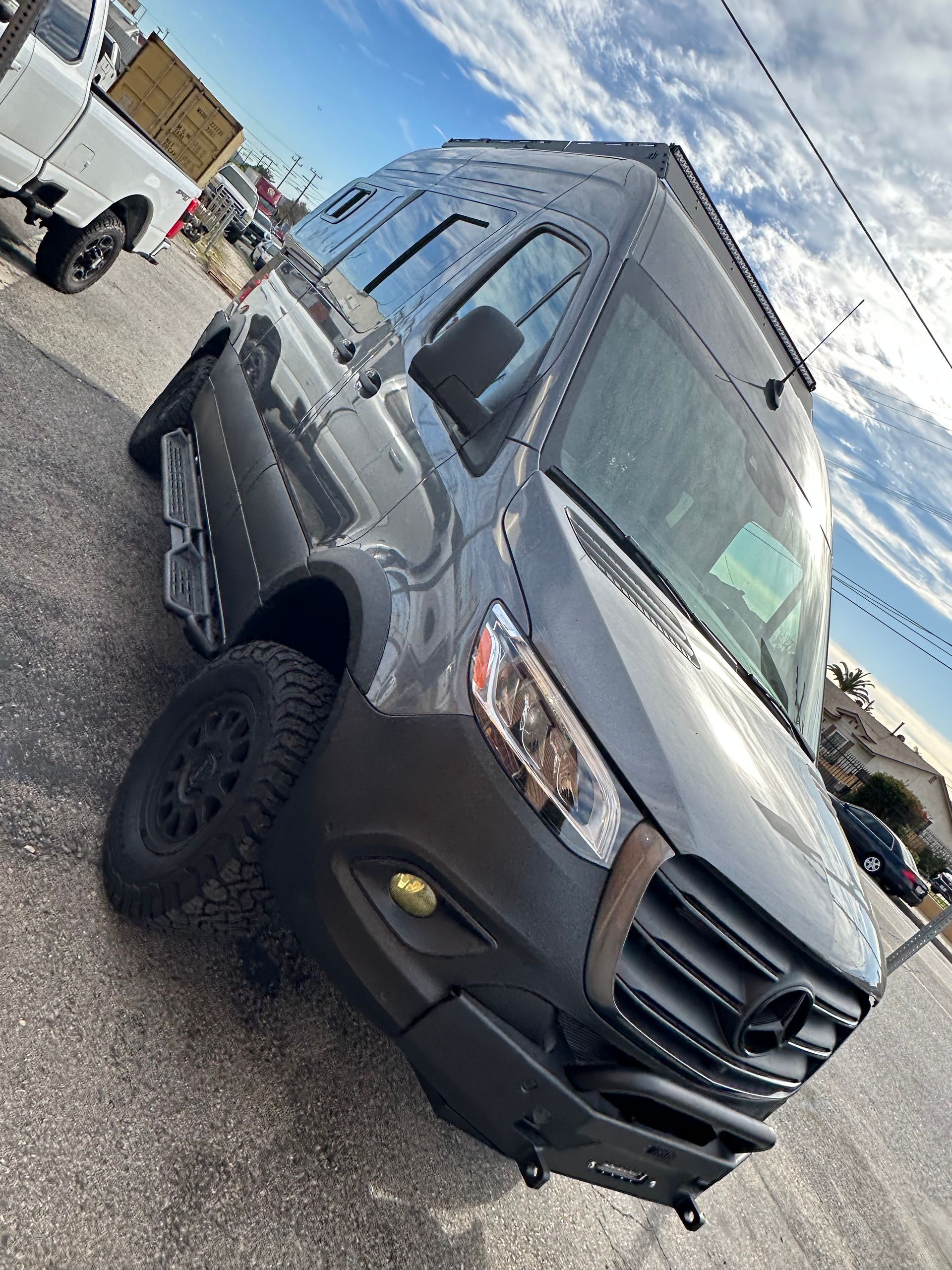 A dark gray, modified Mercedes-Benz Sprinter van with rugged off-road tires and a custom front bumper parked outdoors.
