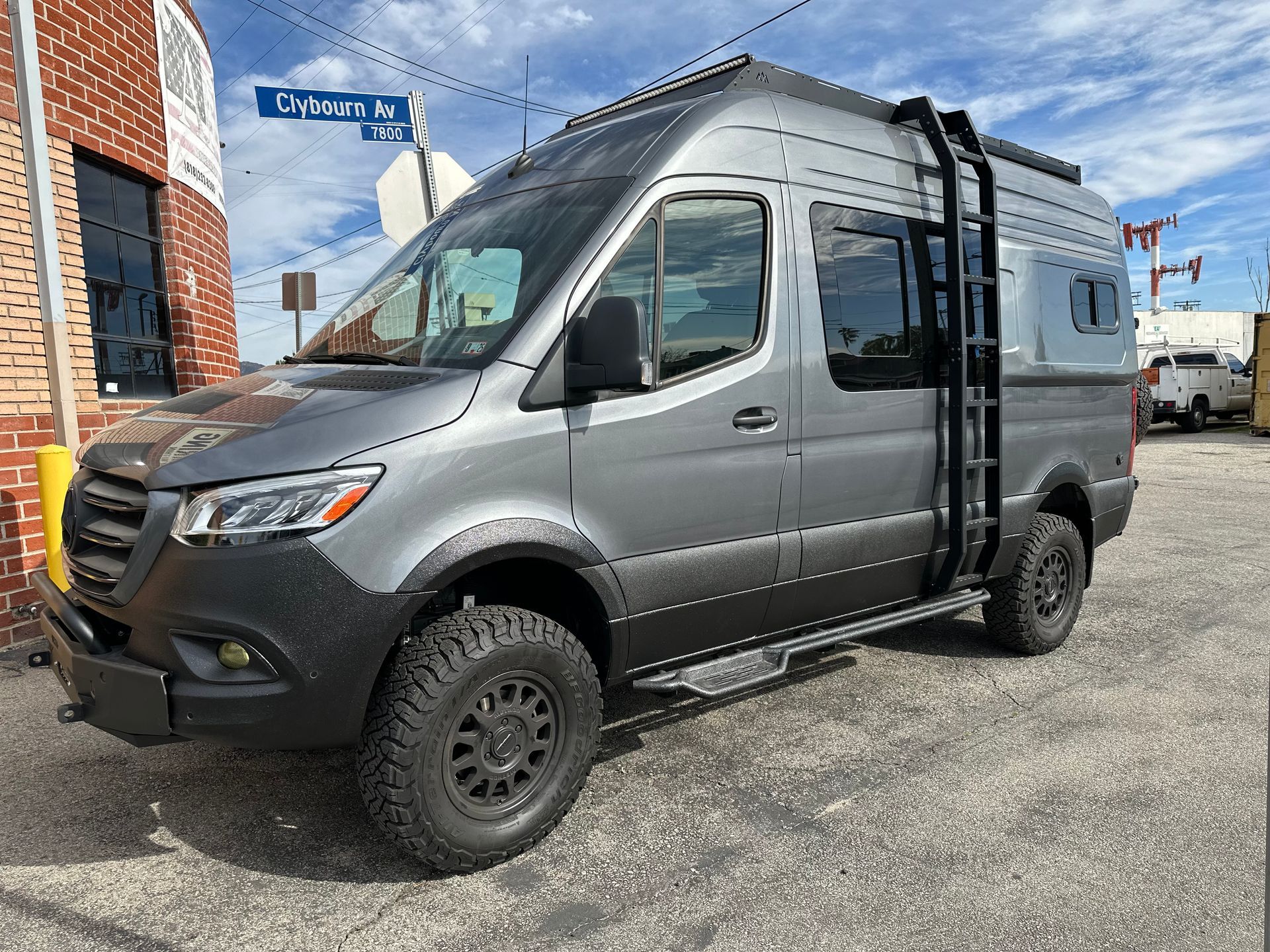 A grey, lifted adventure van with black off-road tires and a side-mounted roof ladder, parked on a street.