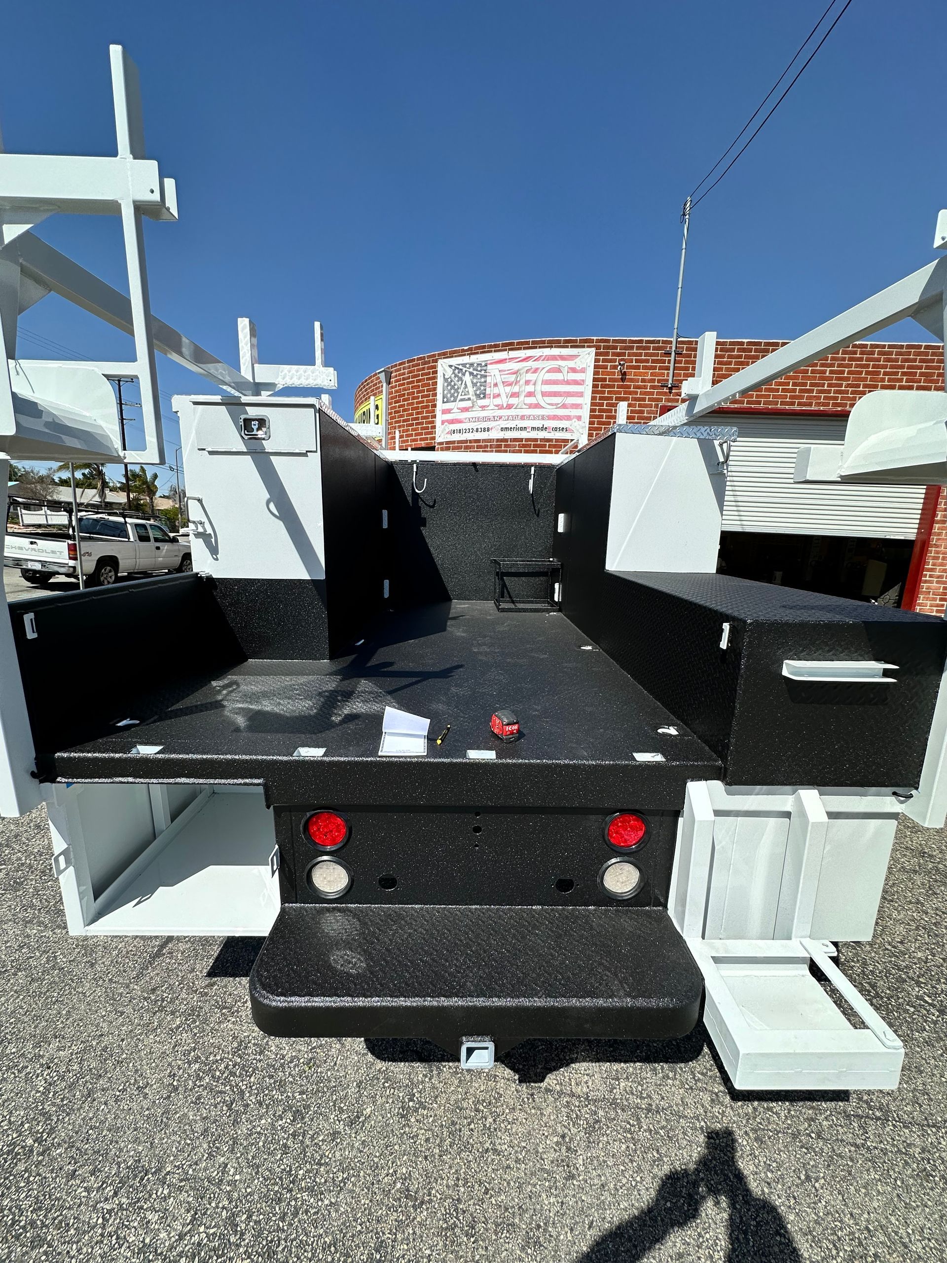 The rear view of a white truck utility bed with black textured coating on the deck, side compartments, and bumper.