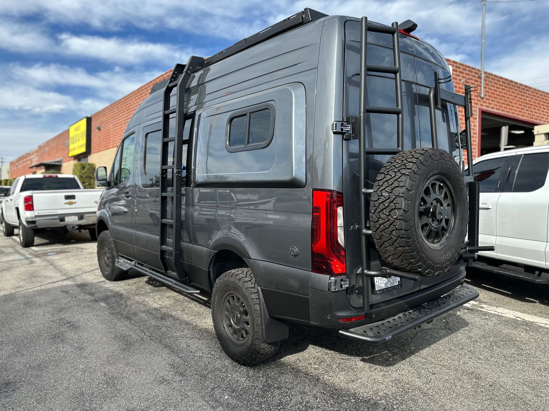A grey camper van with all-terrain tires, a roof rack, and a rear ladder parked in a lot under a blue sky.