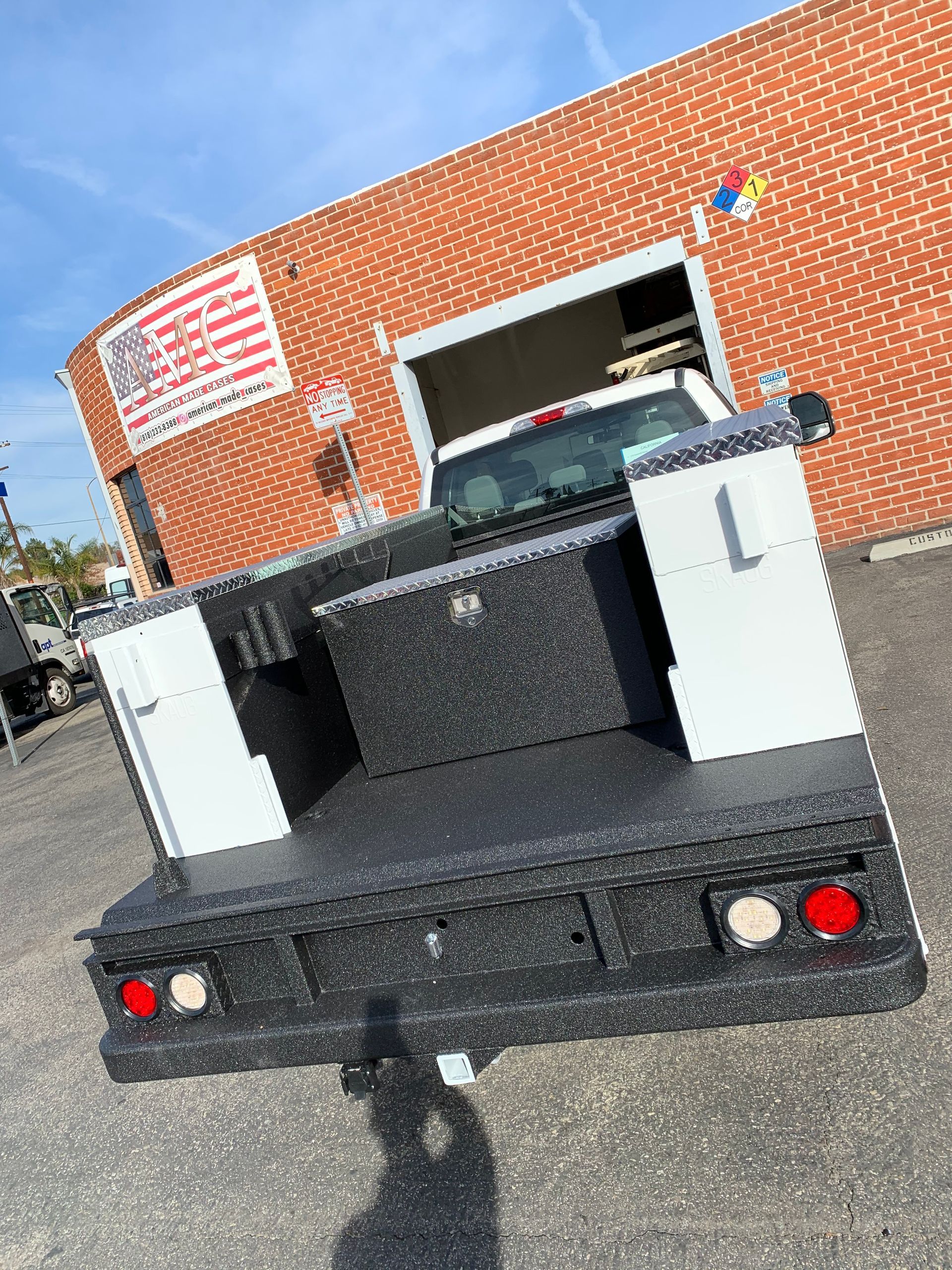 A white pickup truck with a black textured bed liner and metal side toolboxes parked in front of a brick building.