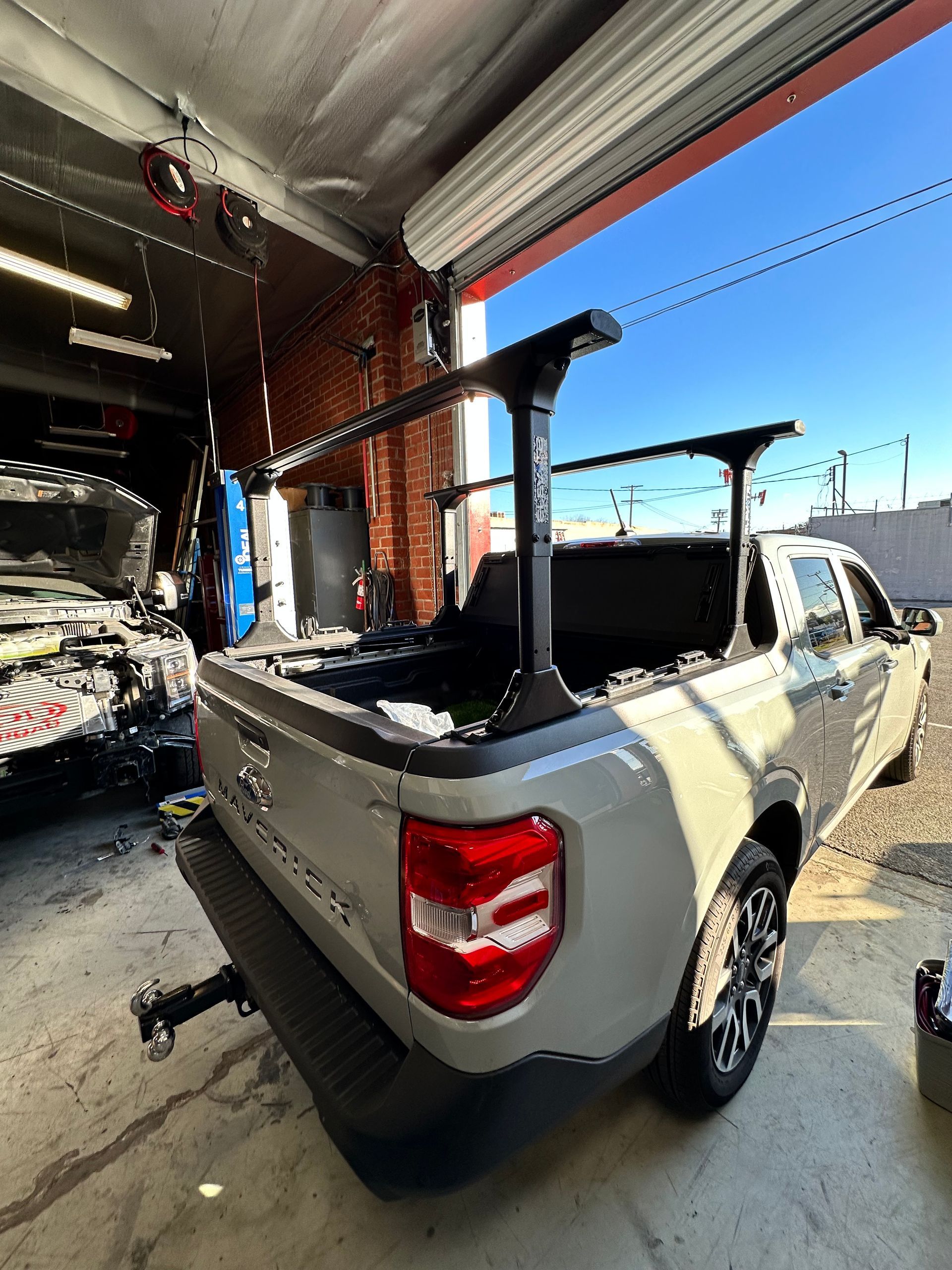 A light grey Ford Maverick pickup truck with a black bed rack installed, parked inside a brightly lit auto shop.