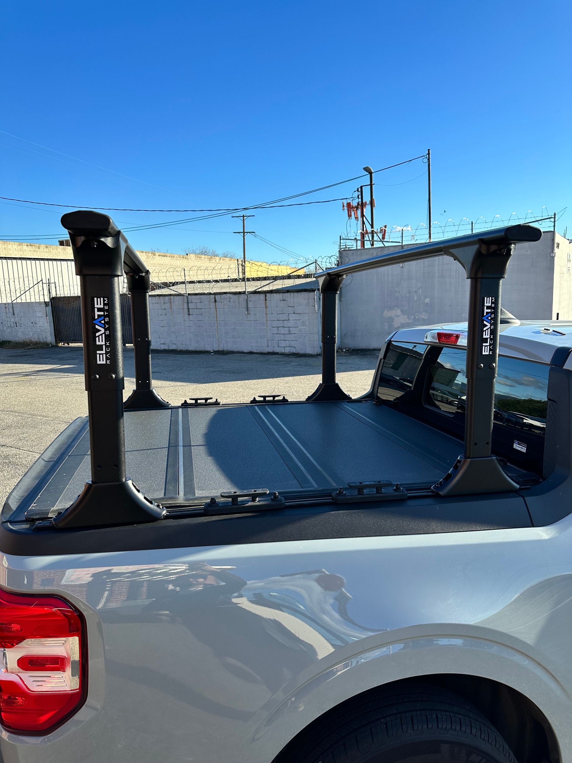 Black truck bed rack mounted on a silver pickup truck bed with a folding cover, parked outdoors under a blue sky.