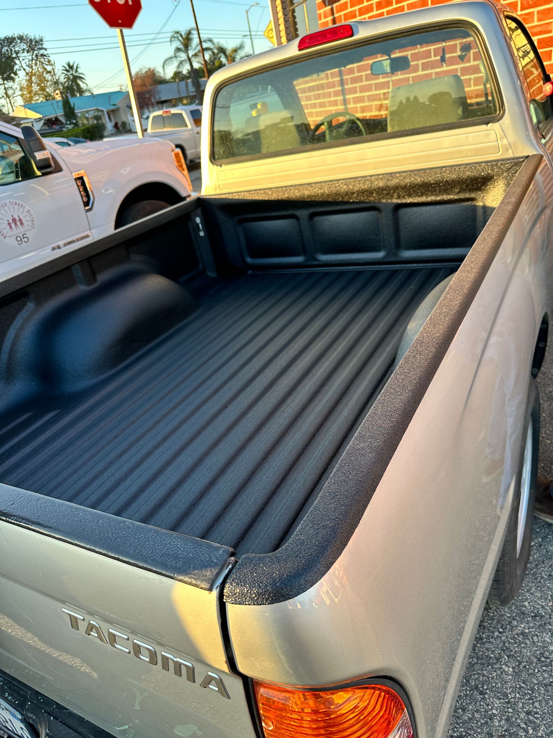 A silver Toyota Tacoma pickup truck, viewed from the rear, showing a black spray-in bed liner in a parking lot.