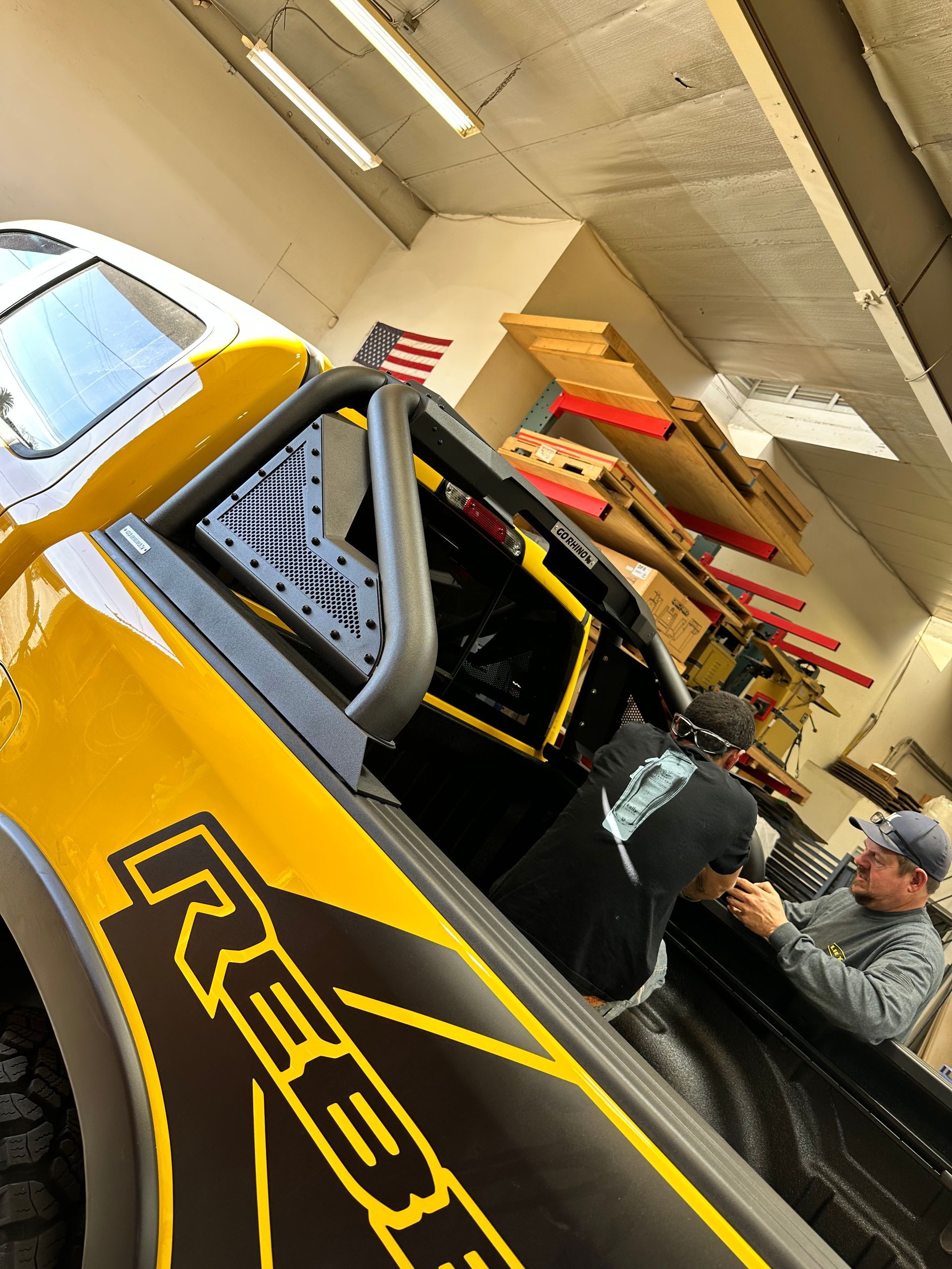 Two individuals work on the interior of a yellow truck featuring a black 