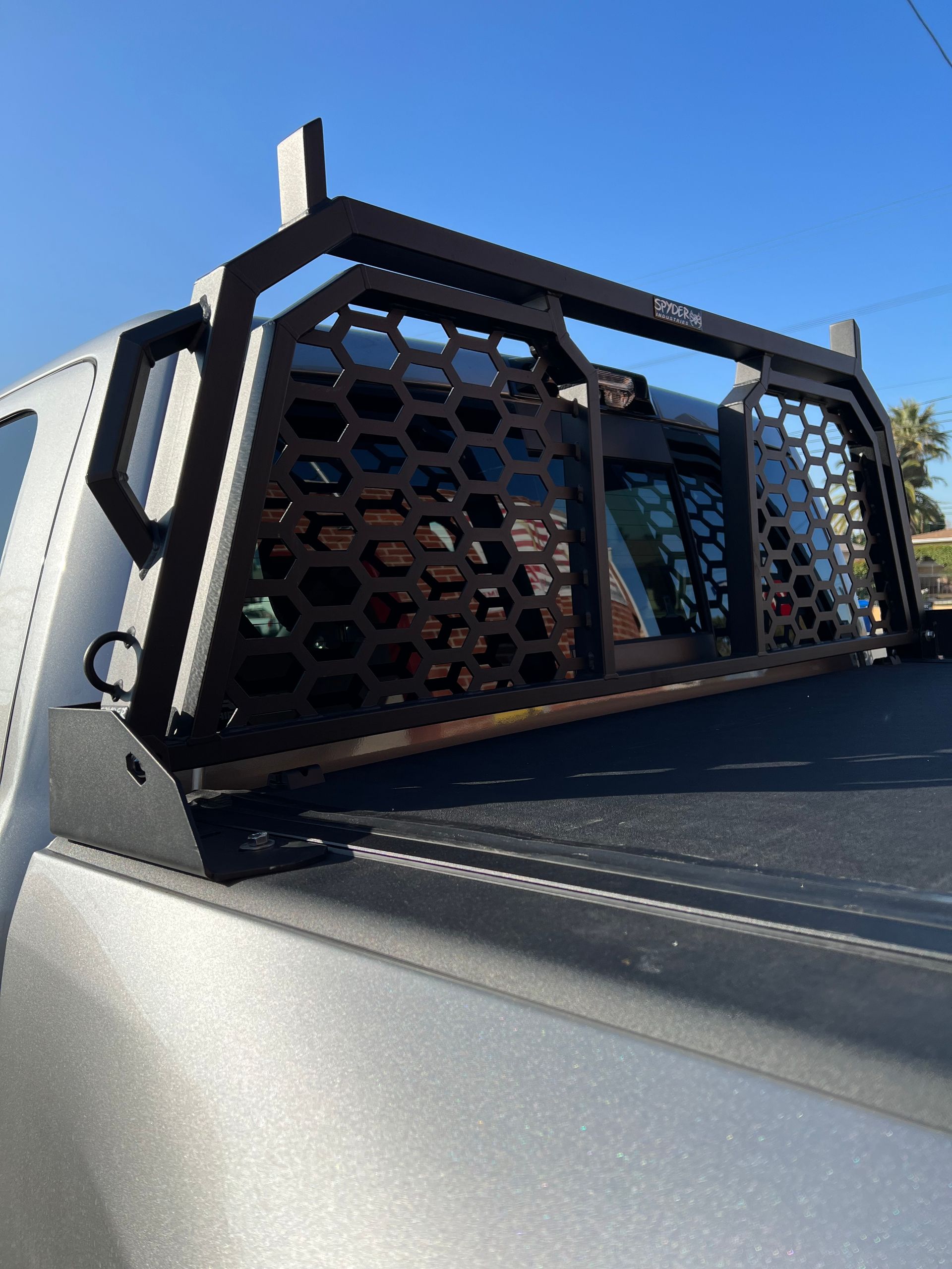Black metal headache rack with a honeycomb mesh design mounted on the bed of a silver truck against a clear blue sky.