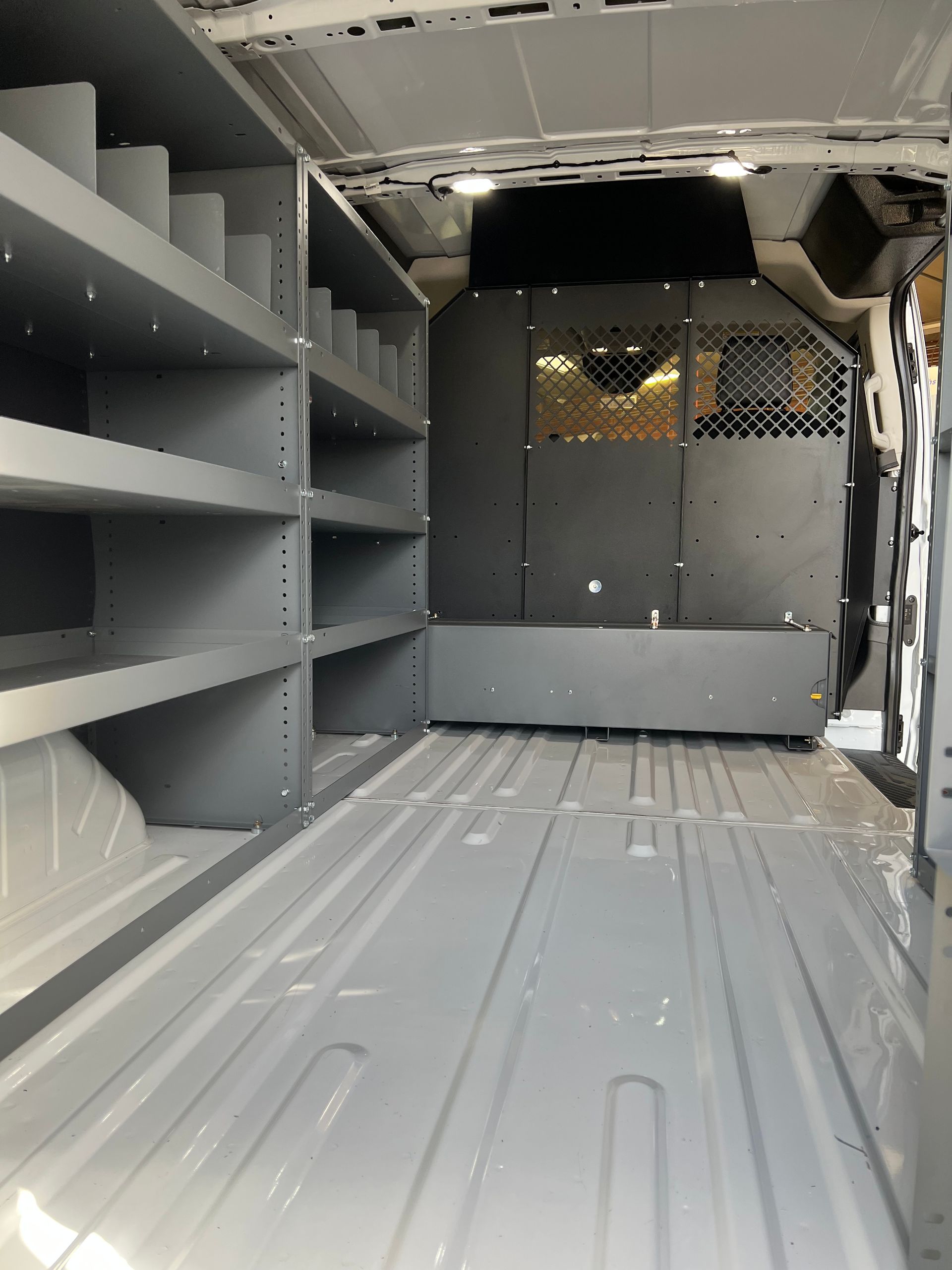 An empty cargo van interior featuring gray metal shelving units on the left and a steel safety partition with mesh.