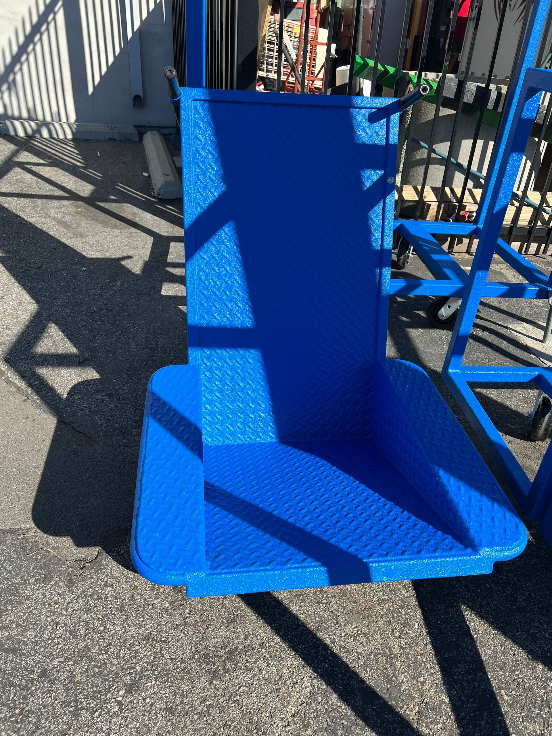 A blue, metal, high-backed swing seat with a diamond-plate texture, suspended outdoors against a metal frame.