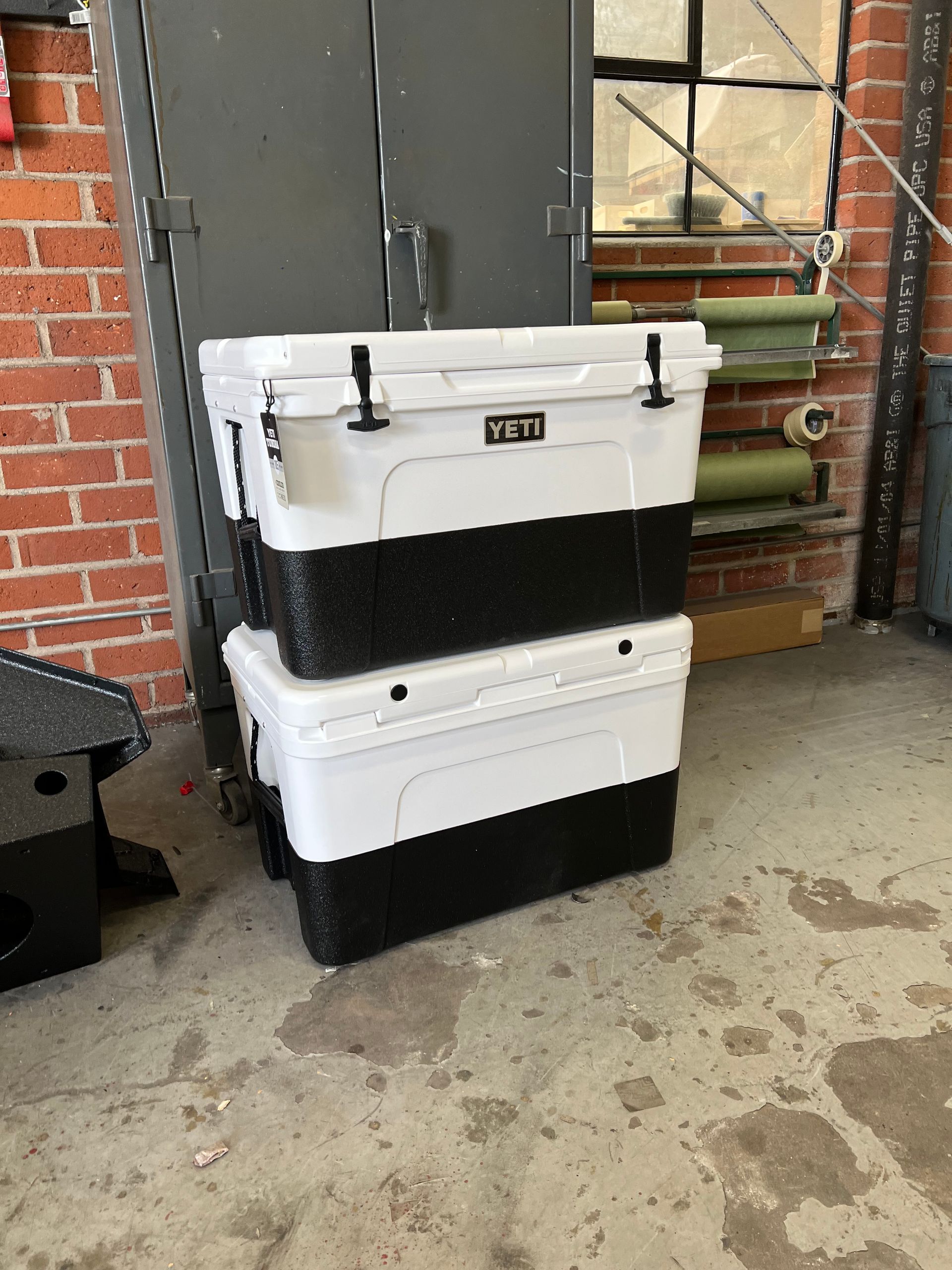 Two stacked, rectangular YETI brand coolers with white tops and black bases sitting on a concrete floor by a brick wall.
