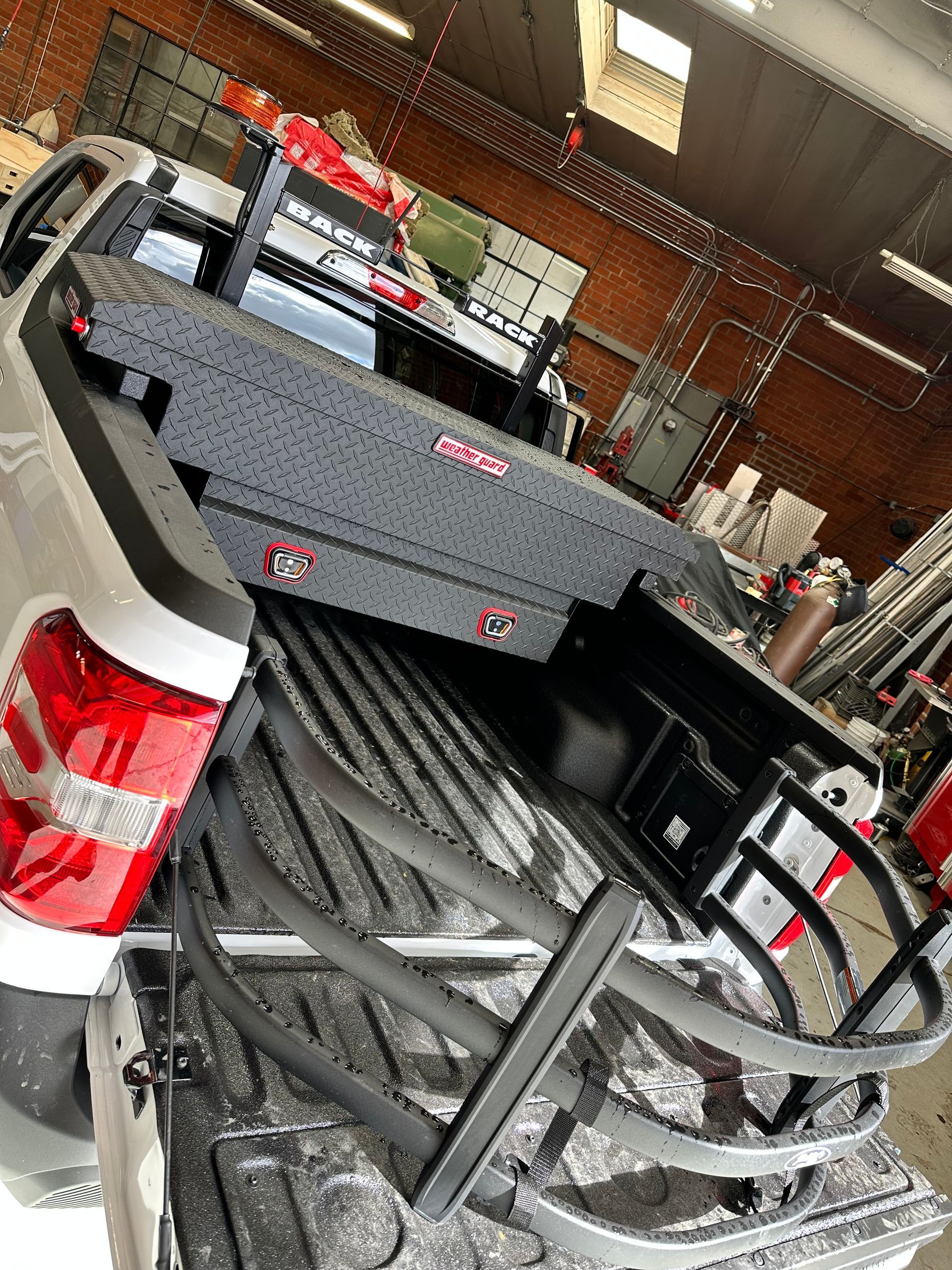A silver pickup truck bed featuring a gray toolbox and a black metal folding bed extender inside a workshop.
