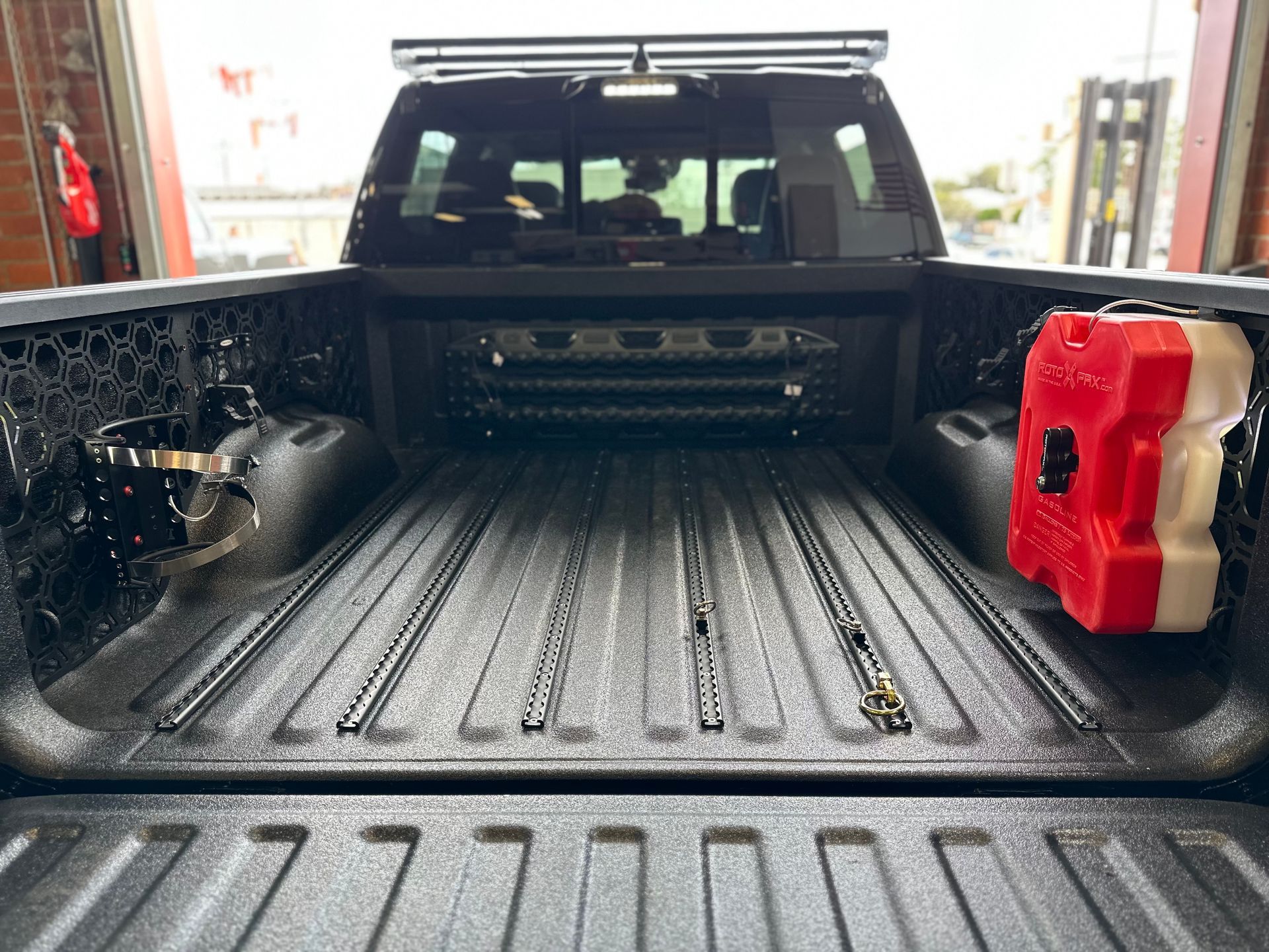 The interior of a black pickup truck bed, featuring floor cargo rails, side wall MOLLE panels, and a red fuel container.