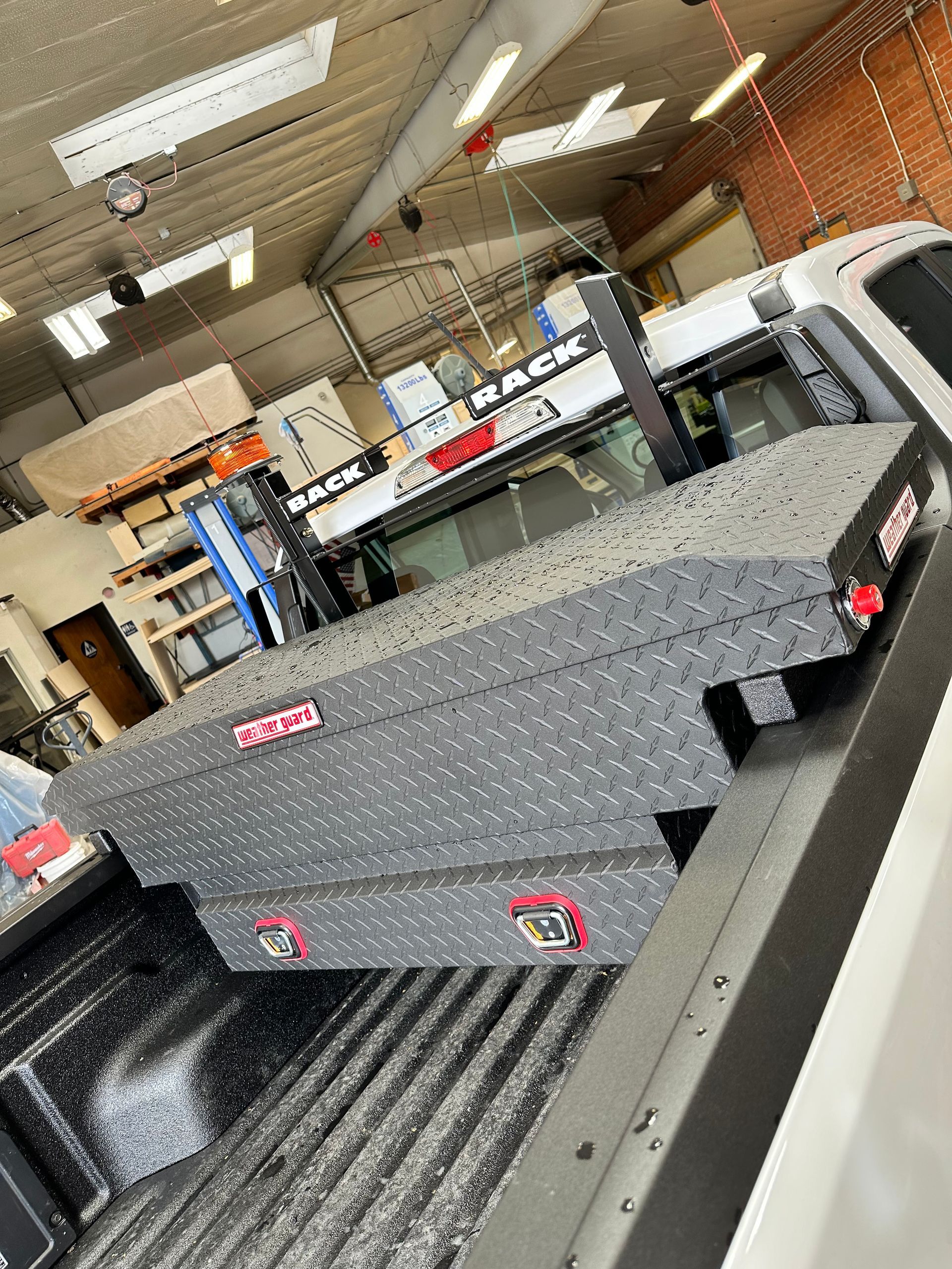 A grey, diamond-plate truck tool box mounted in the bed of a white pickup truck inside a commercial garage.