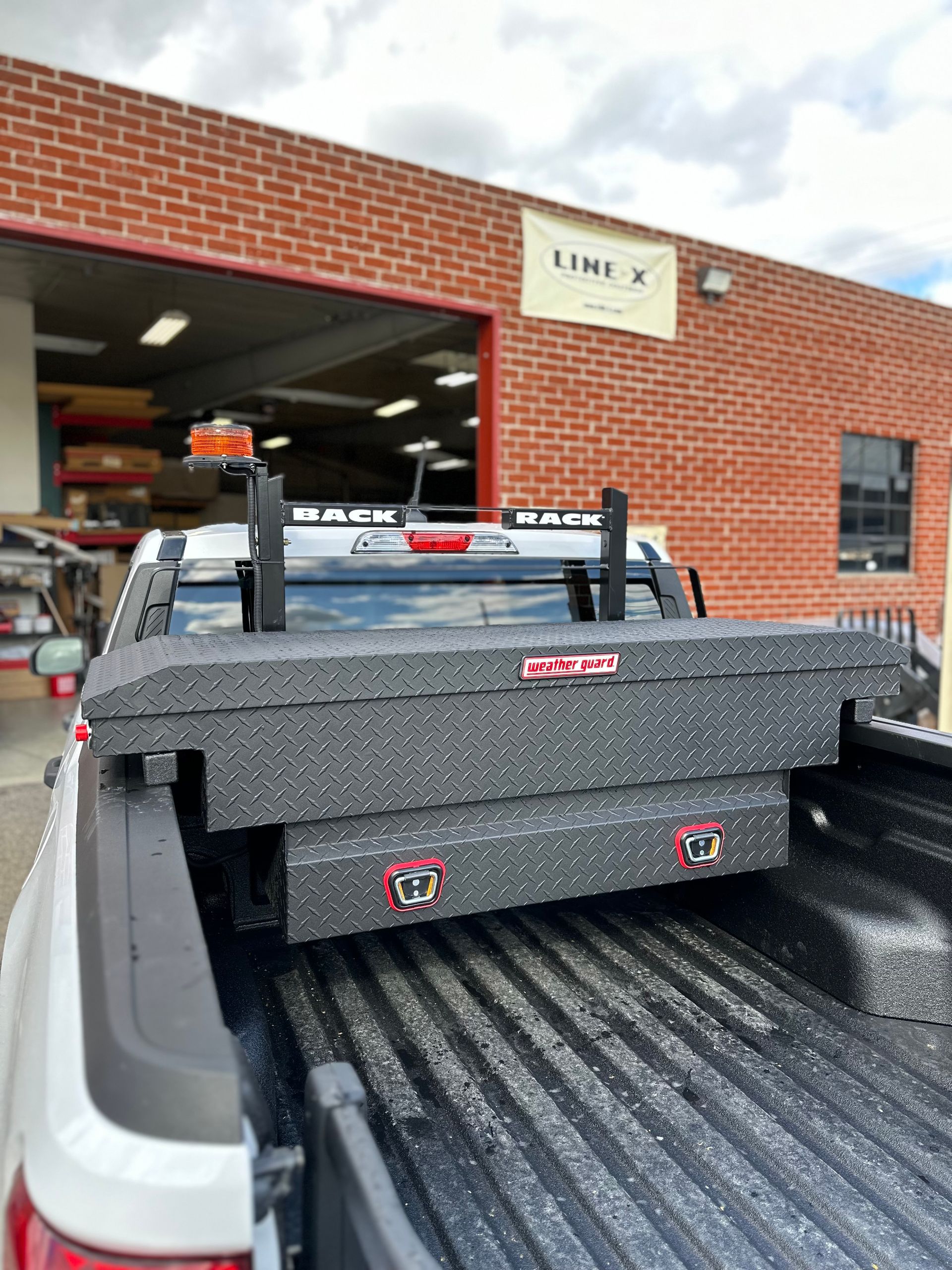 A black metal toolbox with a diamond-plate pattern mounted in the bed of a white pickup truck parked at a business.